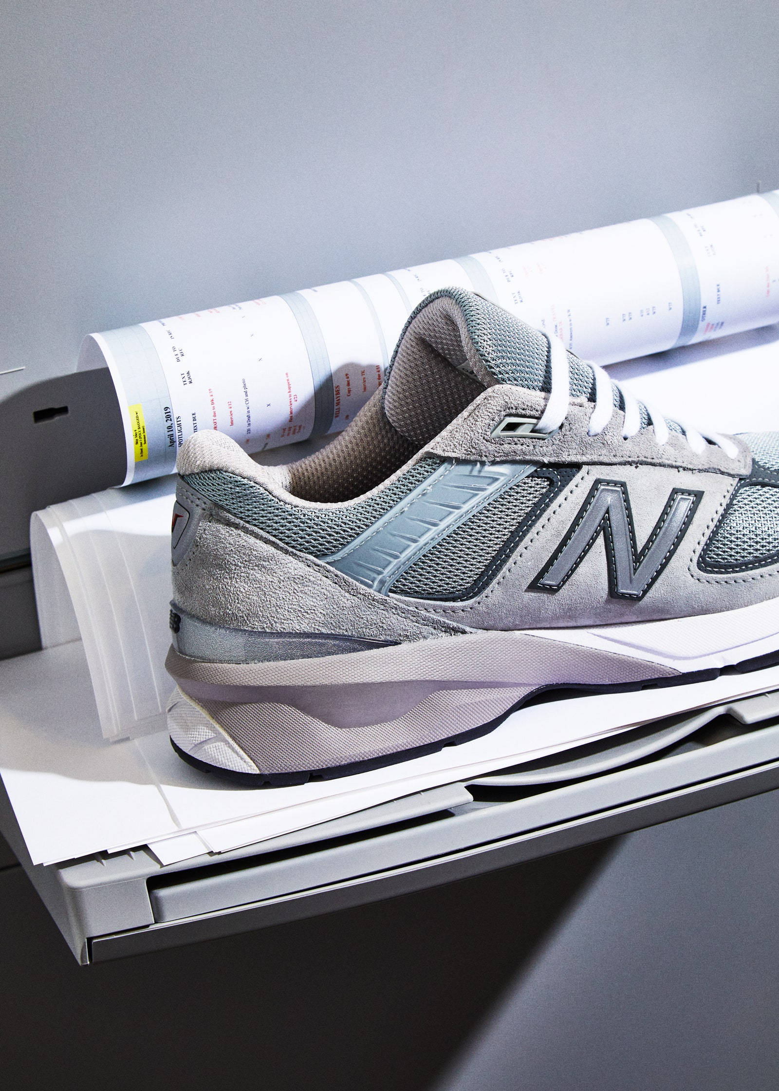 New 桌布 壁紙 — 一雙灰色New Balance運動鞋放在印表機托盤上，旁邊有捲起的技術藍圖，在柔和的工作室燈光下。