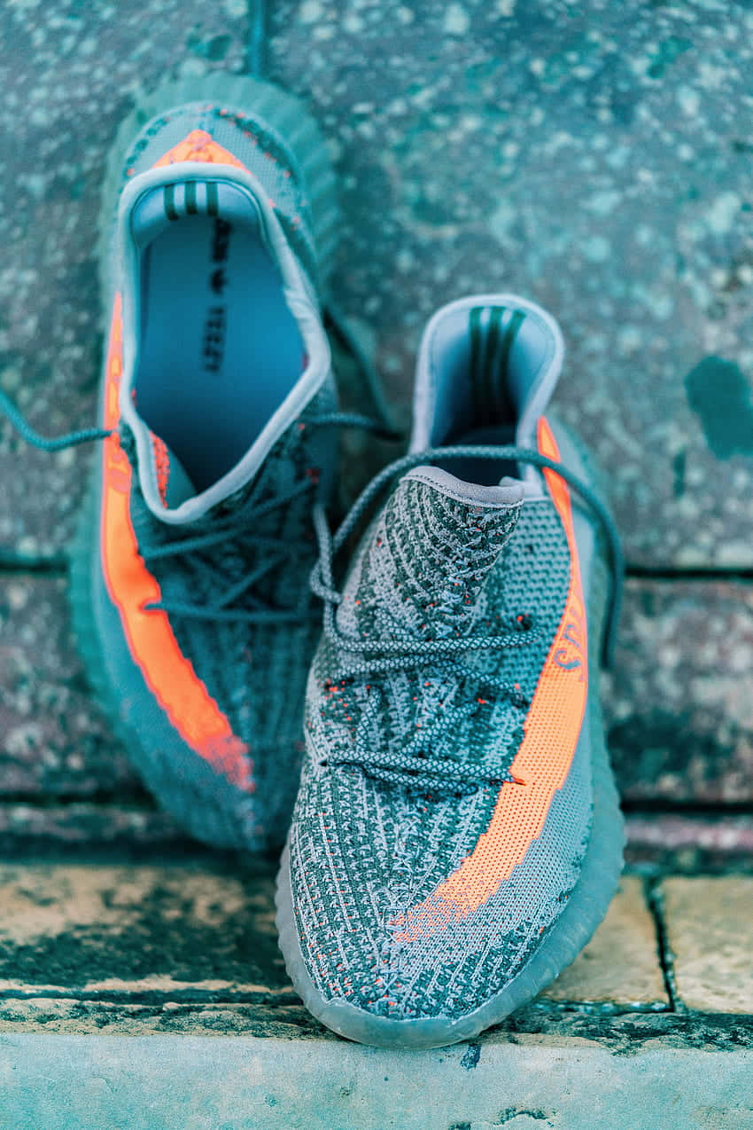Yeezy 桌布 壁紙 — 一雙灰色針織運動鞋帶有橙色點綴，放置在風化的石磚上，背景是紋理牆壁。