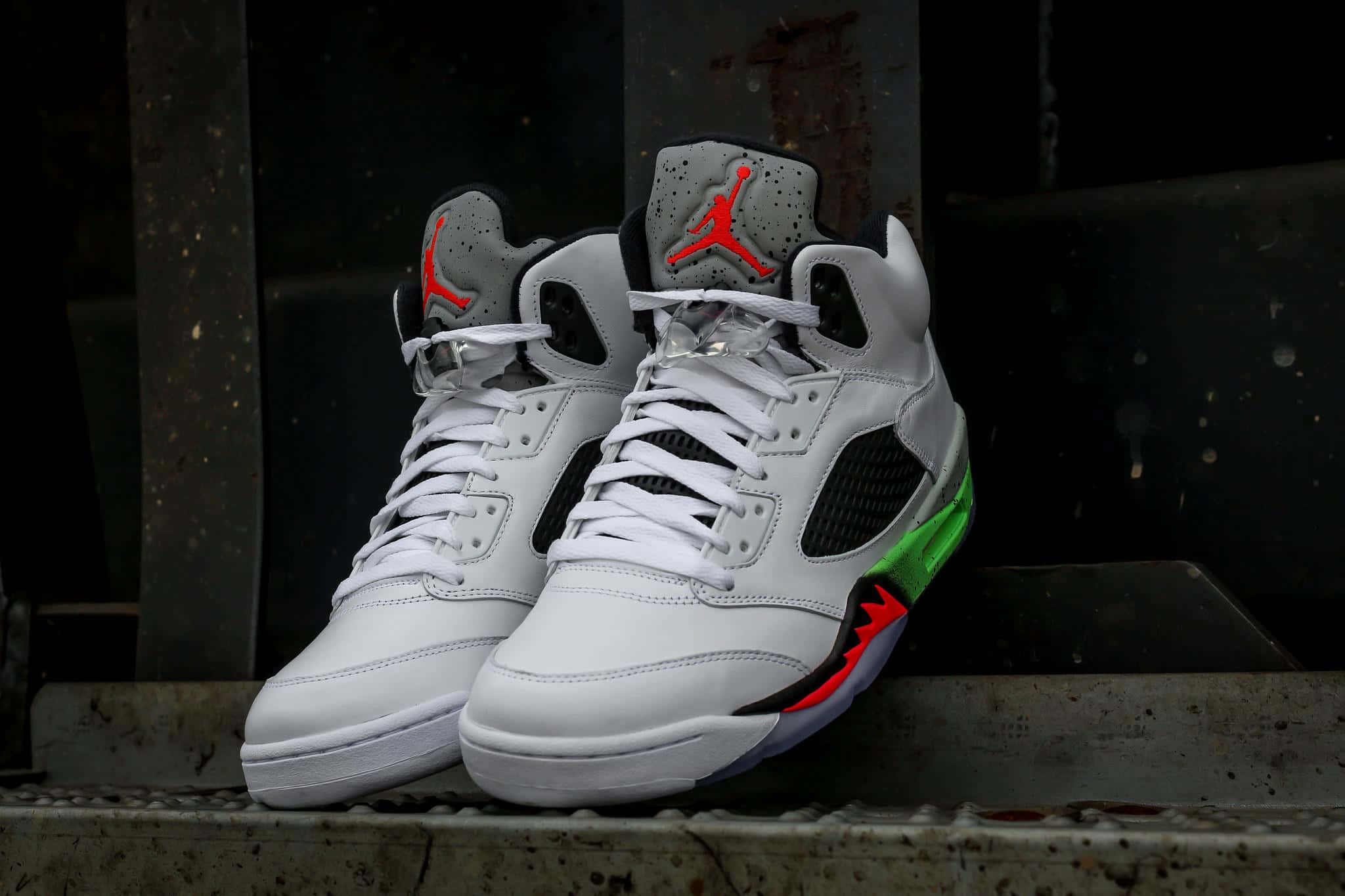 Jordan球鞋 桌布 壁紙 — 一雙白色Jordan 5運動鞋，帶有紅色和綠色點綴，放在風化的混凝土邊緣上，背景是黑暗的工業環境。