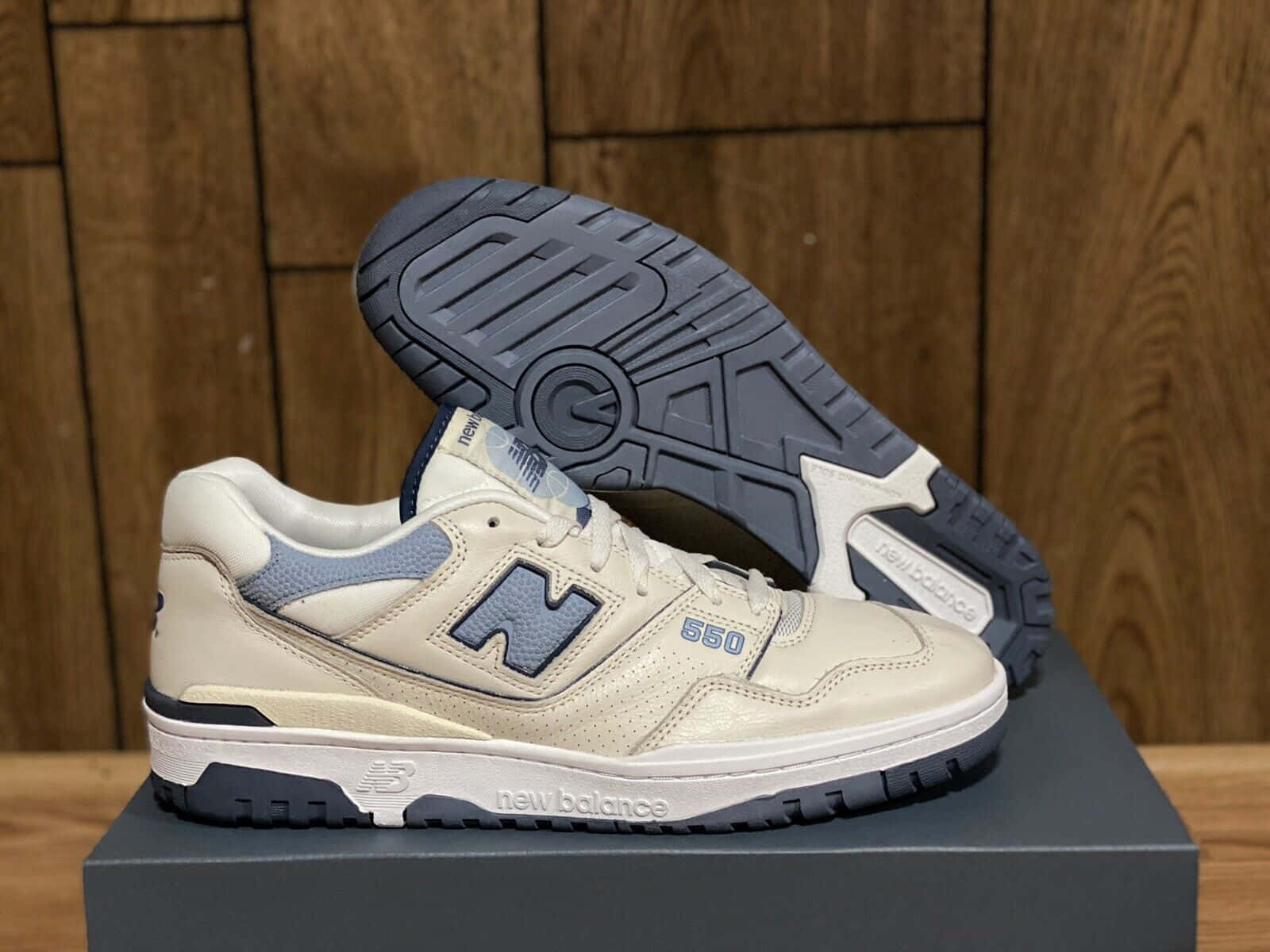New Balance 桌布 壁紙 — 一雙米色和海軍藍色的New Balance 550運動鞋展示在鞋盒上，鞋底翹起，背景為木牆。