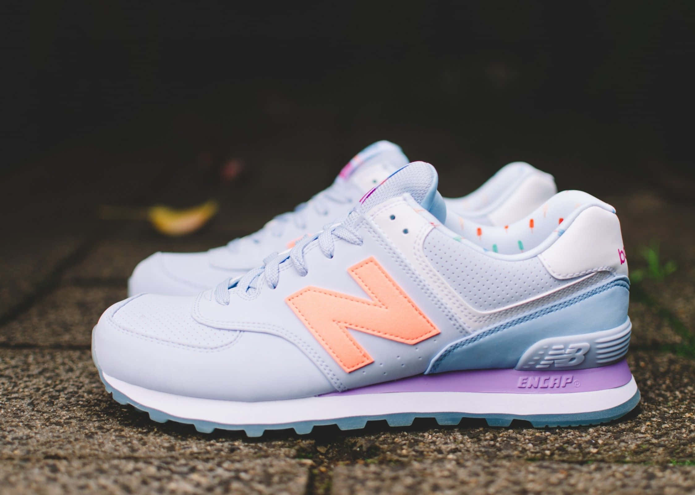 New Balance 桌布 壁紙 — 一雙粉白色New Balance運動鞋，帶有蜜桃橙色N標誌和紫色鞋底，放在石鋪地面上。