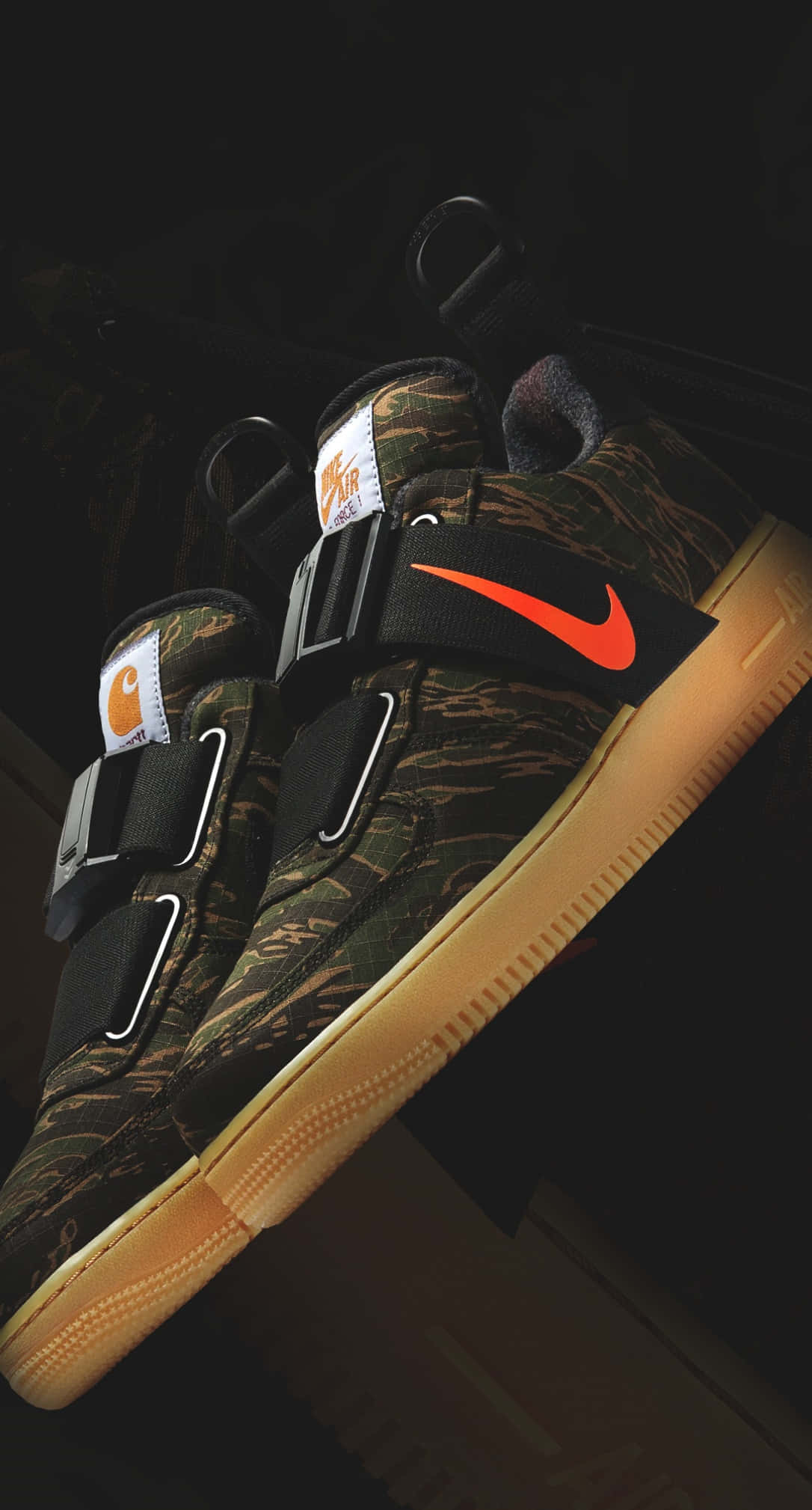 桌布 壁紙 — 一雙迷彩 Nike Carhartt 運動鞋，帶有醒目的橙色 swoosh 標誌和棕褐色鞋底，在深色背景下戲劇性地照亮。
