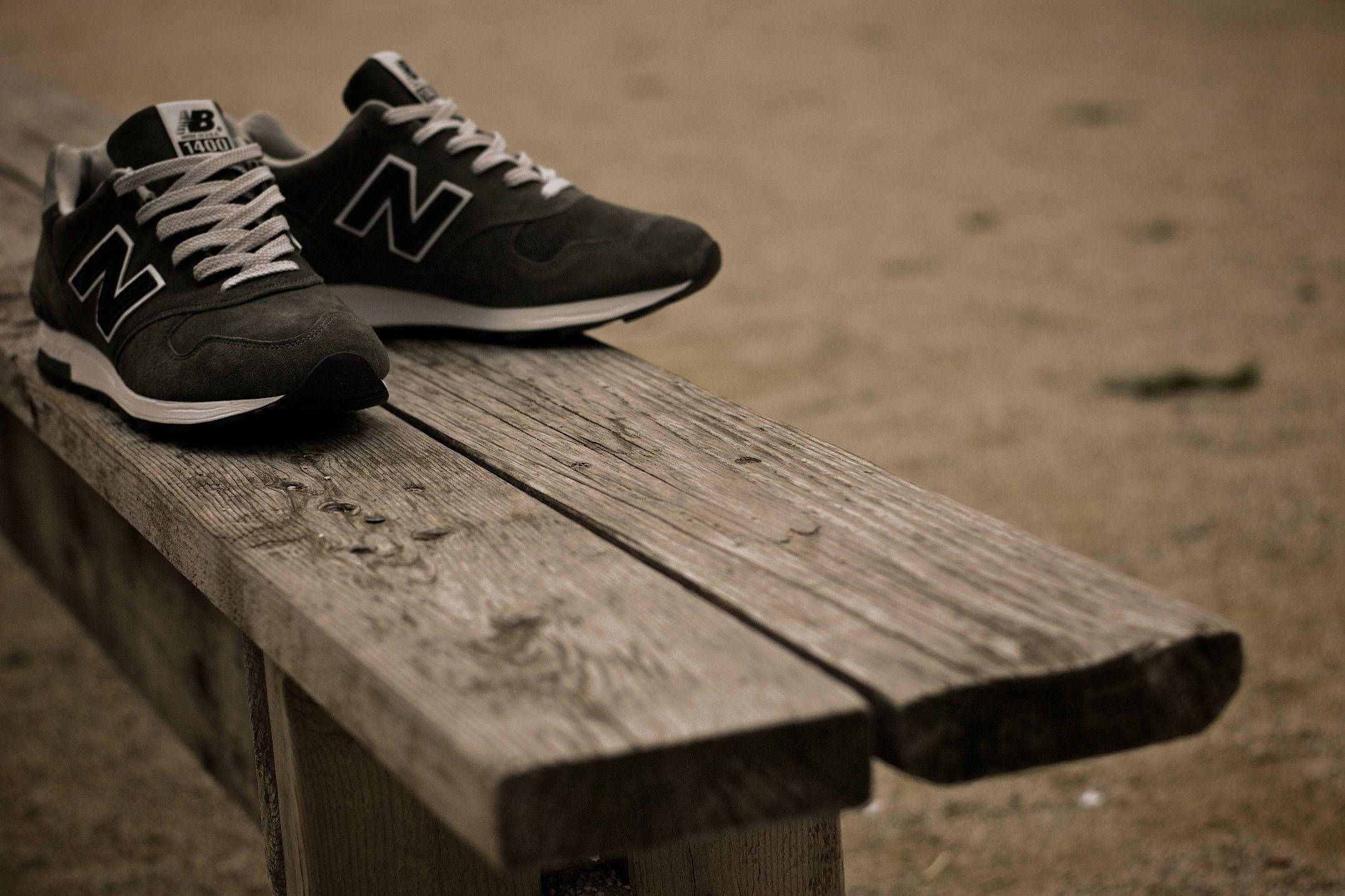 New 桌布 壁紙 — 一雙黑色New Balance運動鞋搭配白色鞋帶，擱置在一張風化的木長椅上，背景是沙地。