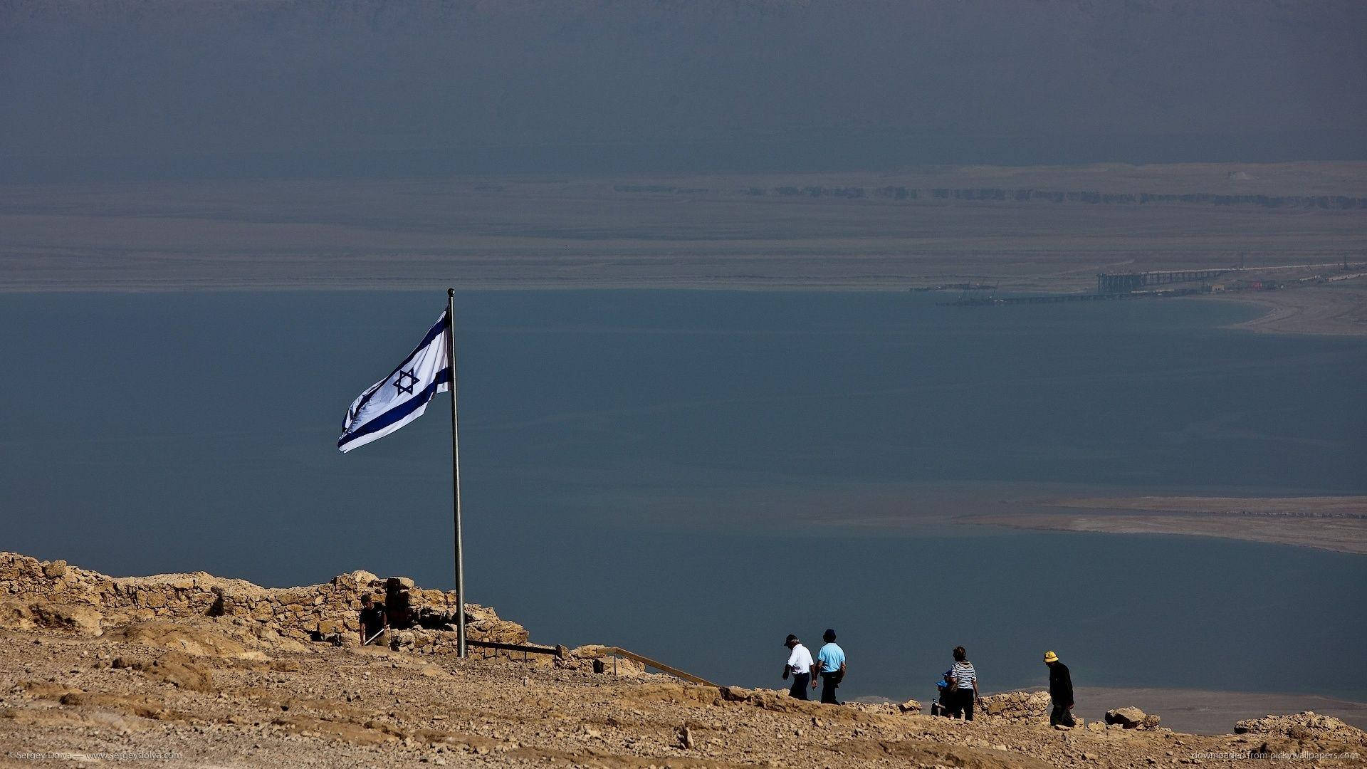 Israel Flag 桌布 壁紙 — 一面以色列國旗在岩石山頂上飄揚，俯瞰著廣闊的藍色加利利海，遠處有身影在下方行走。