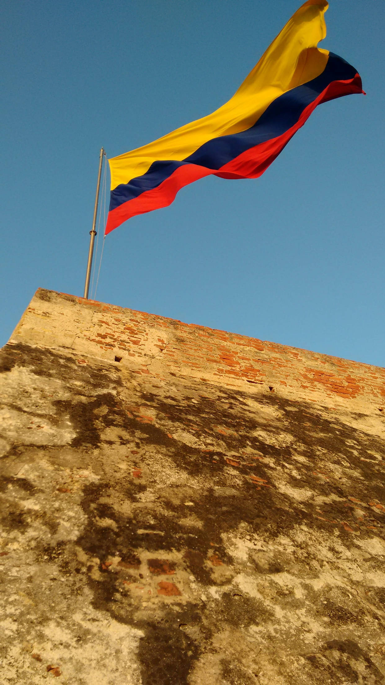 Colombia Flag 桌布 壁紙 — 一面充滿活力的哥倫比亞國旗在歷經風霜的磚砌堡壘頂部驕傲地飄揚，映襯著清澈的藍天。