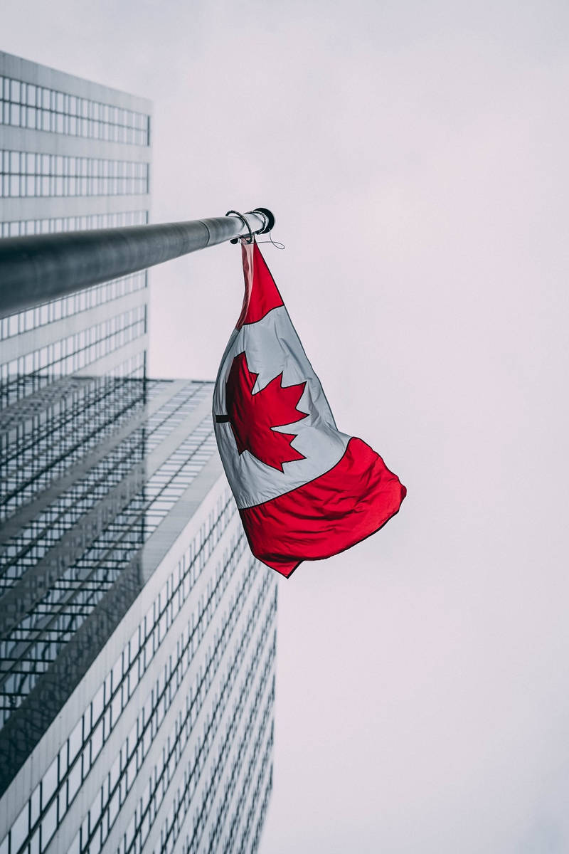Canada Flag 桌布 壁紙 — 一面加拿大國旗在陰天背景下，無力地垂掛在旗桿上，背景是一座現代玻璃摩天大樓。