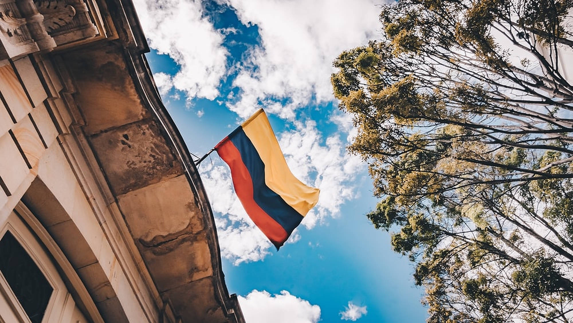 Colombia 桌布 壁紙 — 一面哥倫比亞國旗在晴朗藍天和 fluffy 白雲襯托下驕傲飄揚，周圍有華麗建築和茂密樹枝框景。