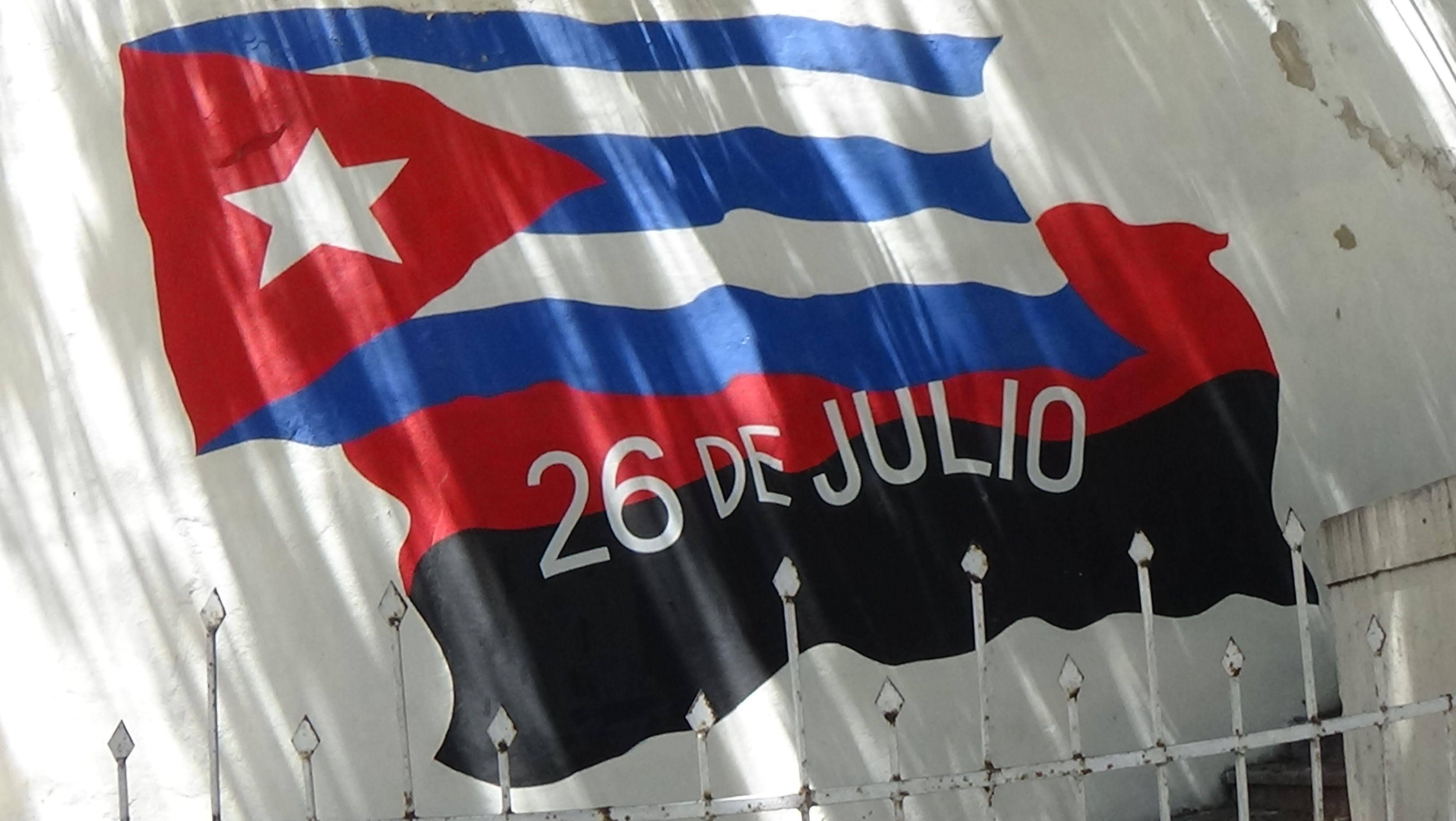 Cuban 桌布 壁紙 — 一面大型古巴國旗，上面繪有「26 DE JULIO」字樣，在生鏽金屬圍欄後方，對著風化的白色牆壁飄揚。