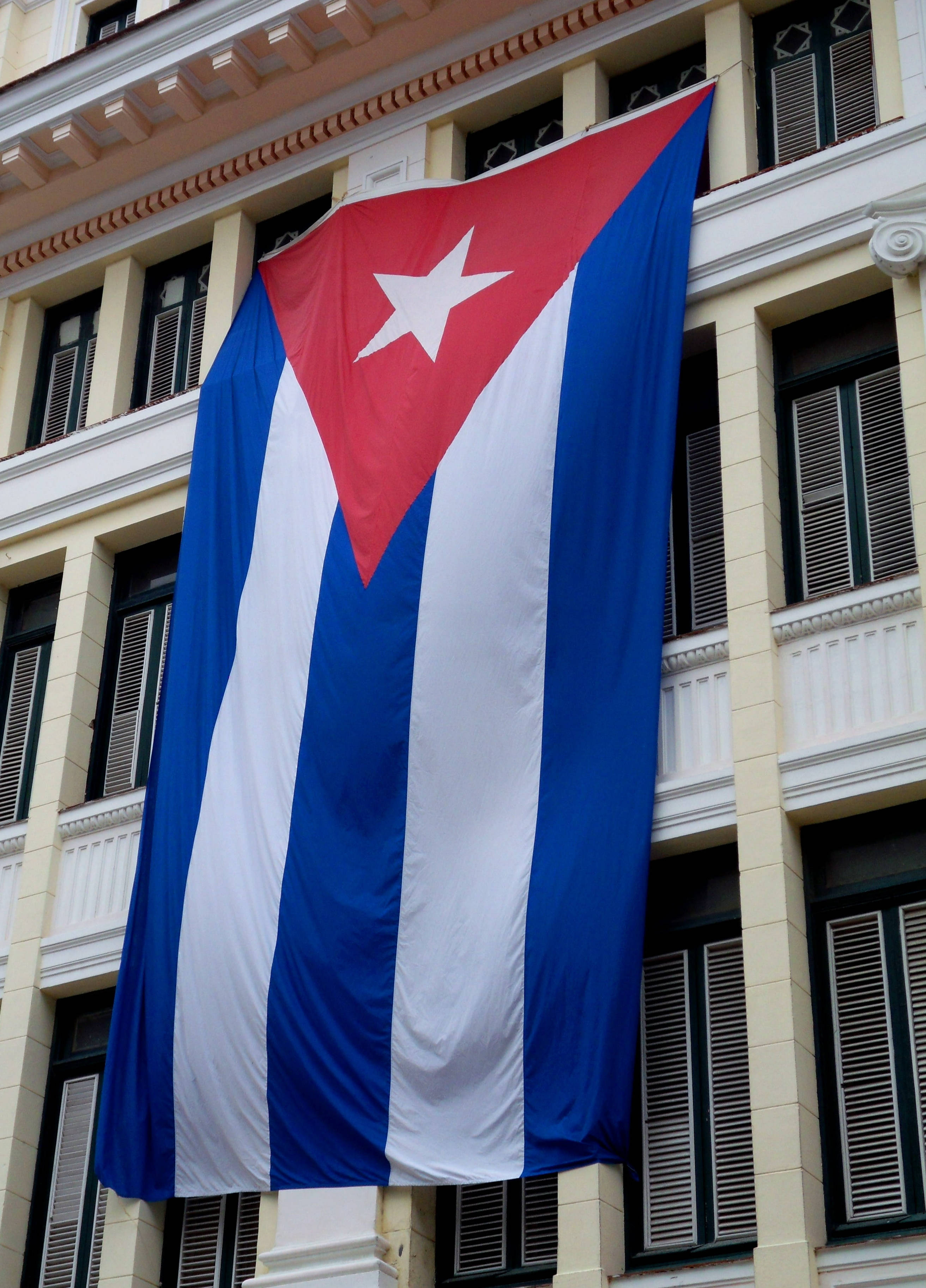 Cuban 桌布 壁紙 — 一面大型古巴國旗，帶有紅、白、藍條紋和一顆白色星星，醒目地懸掛在古典建築的外牆上。