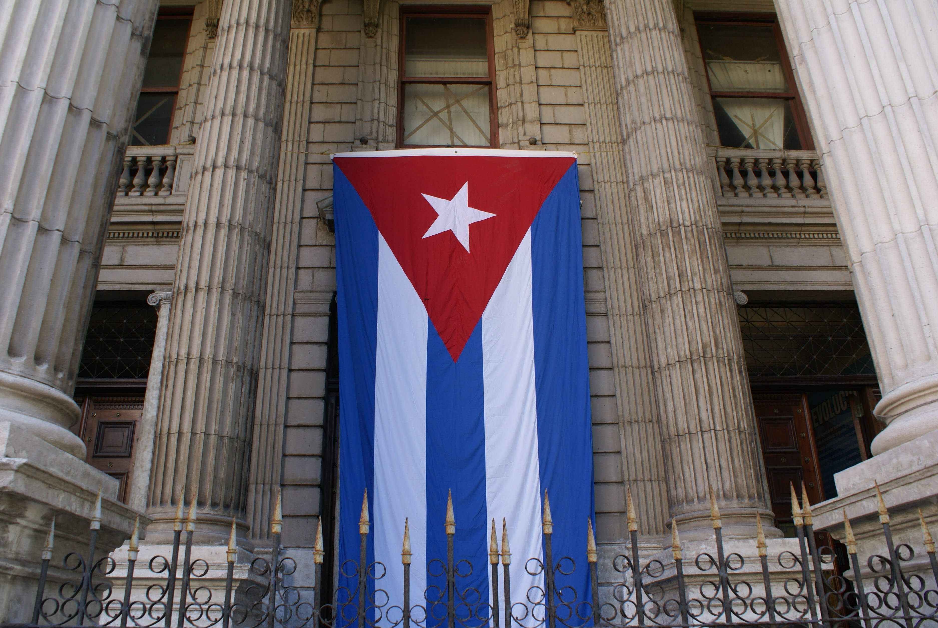Cuban 桌布 壁紙 — 一面大型古巴國旗在日光下顯眼地懸掛於宏偉新古典主義建築的石柱之間。