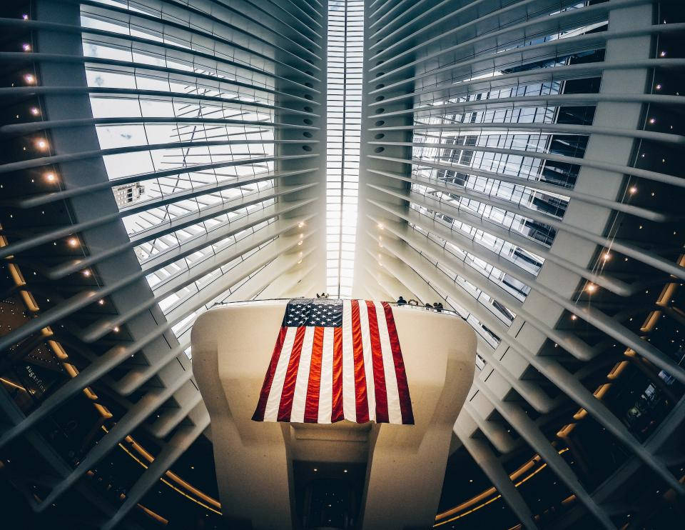 USA flag iPhone 桌布 壁紙 — 一面大型美國國旗莊嚴地懸掛在一個高聳的現代中庭中央，該中庭採用弧形玻璃和鋼結構建築。
