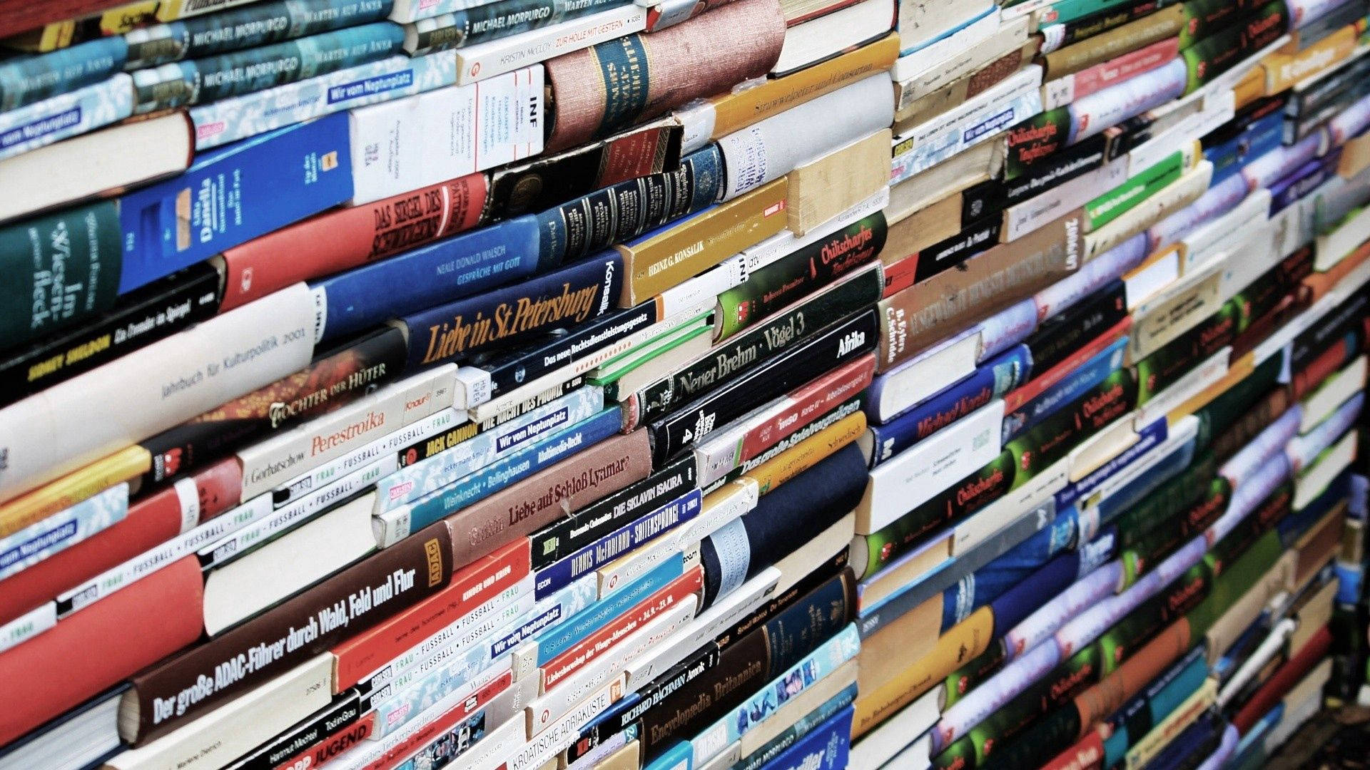 書本 桌布 壁紙 — 一面密集的彩色書牆，書本以傾斜角度緊密堆疊，書脊朝外，形成充滿活力的文學拼貼畫。