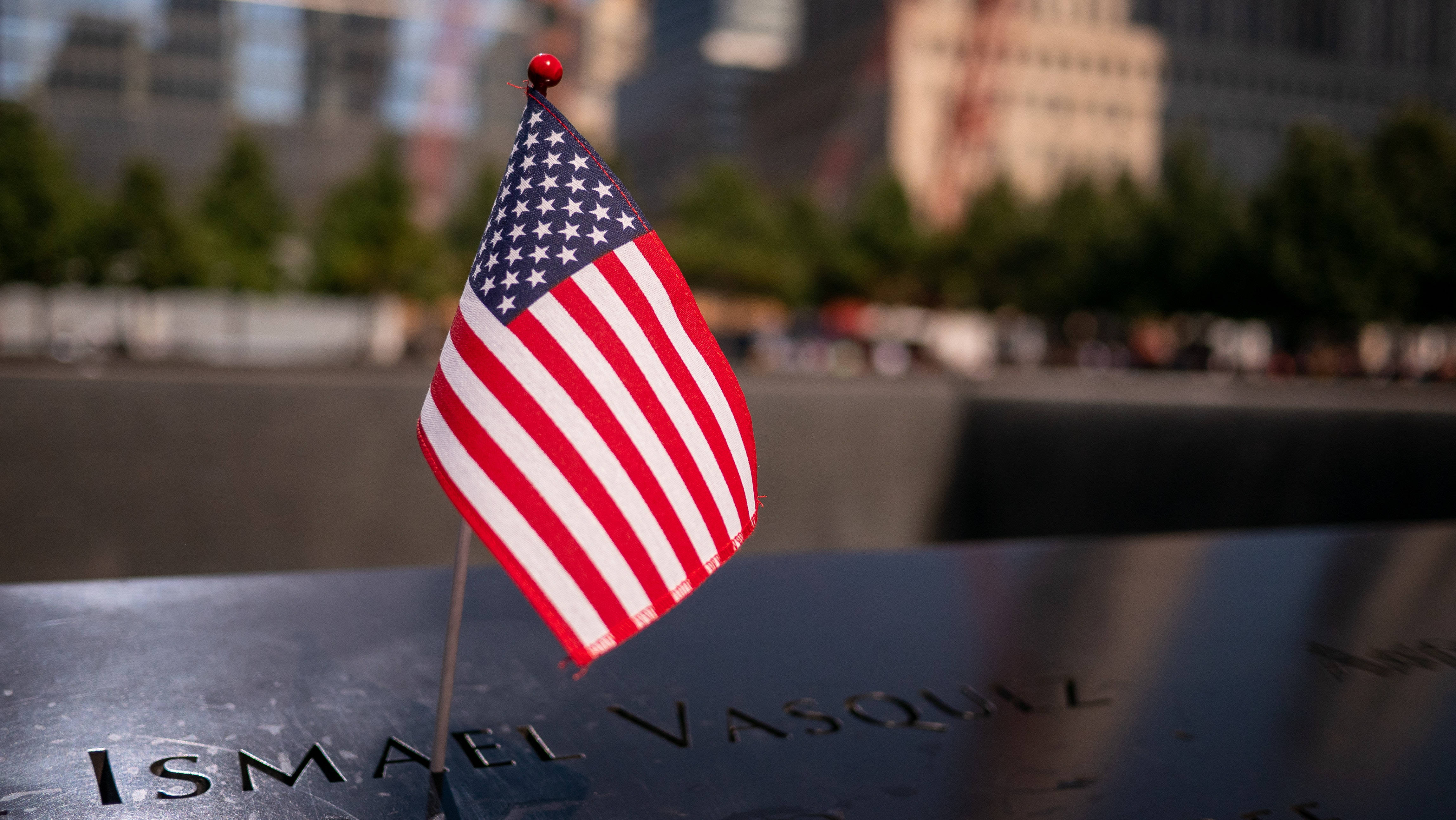 911 Memorial 桌布 壁紙 — 一面小型美國國旗莊嚴地豎立在刻有名字的黑色紀念牌上，背景是模糊的城市景觀。