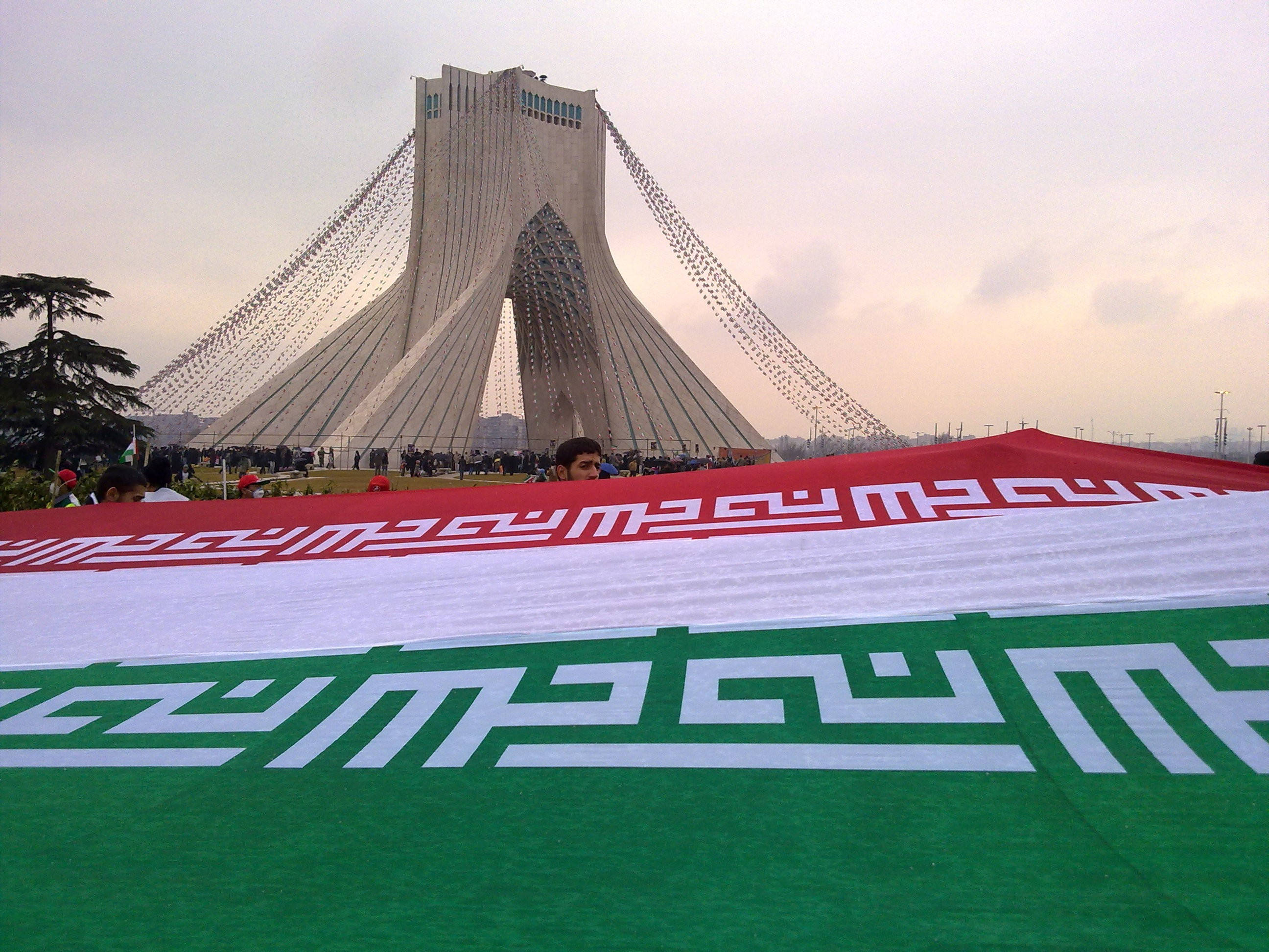 Tehran 桌布 壁紙 — 一面巨大的伊朗國旗在黃昏時分於標誌性的阿扎迪塔前展開，前景聚集著人群。