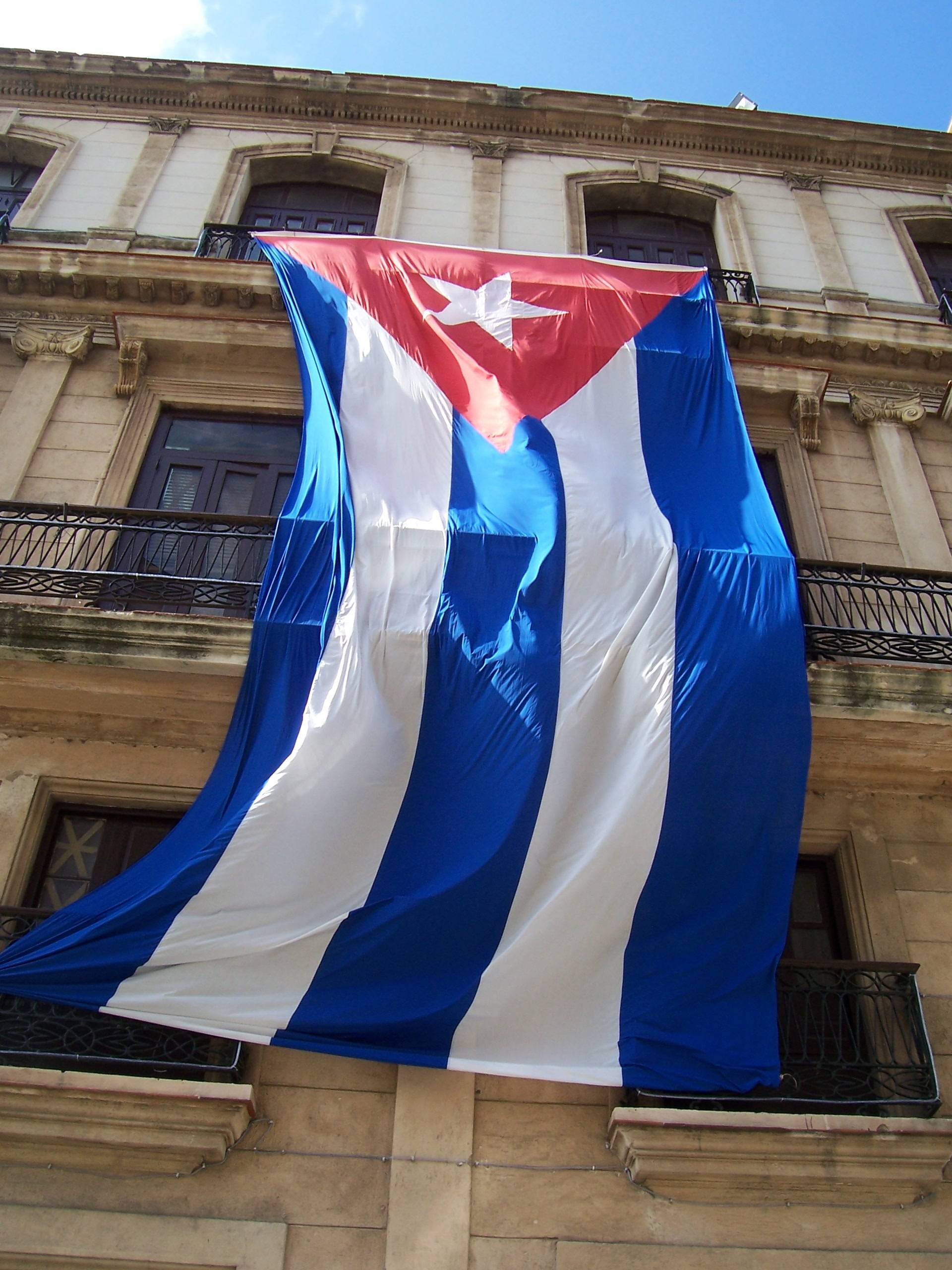 Cuba 桌布 壁紙 — 一面巨大的古巴國旗在晴朗的藍天下，在歷史悠久的石建築前戲劇性地飄揚。