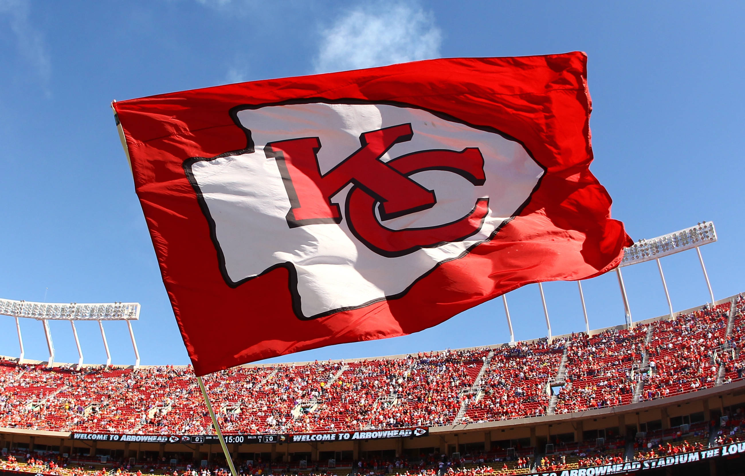 Chiefs Logo 桌布 壁紙 — 一面巨大的紅色堪薩斯城酋長旗幟在晴朗的藍天下，在擠滿歡呼球迷的體育場上空自豪地飄揚。
