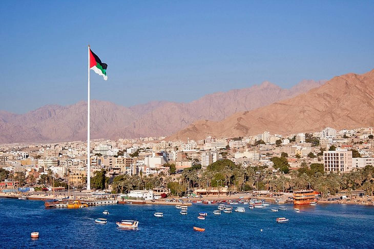 Jordan 桌布 壁紙 — 一面巴勒斯坦國旗在旗桿上驕傲地飄揚，俯瞰著一個沿海城市，藍色海上有船隻，背景是乾旱的山脈。