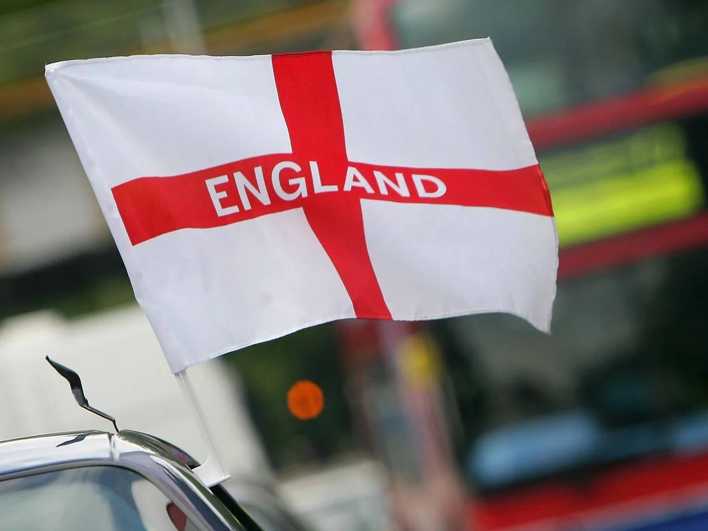 England Flag 桌布 壁紙 — 一面帶有 'ENGLAND' 字樣的紅色十字旗在汽車天線上飄揚，背景是模糊的城市景觀。