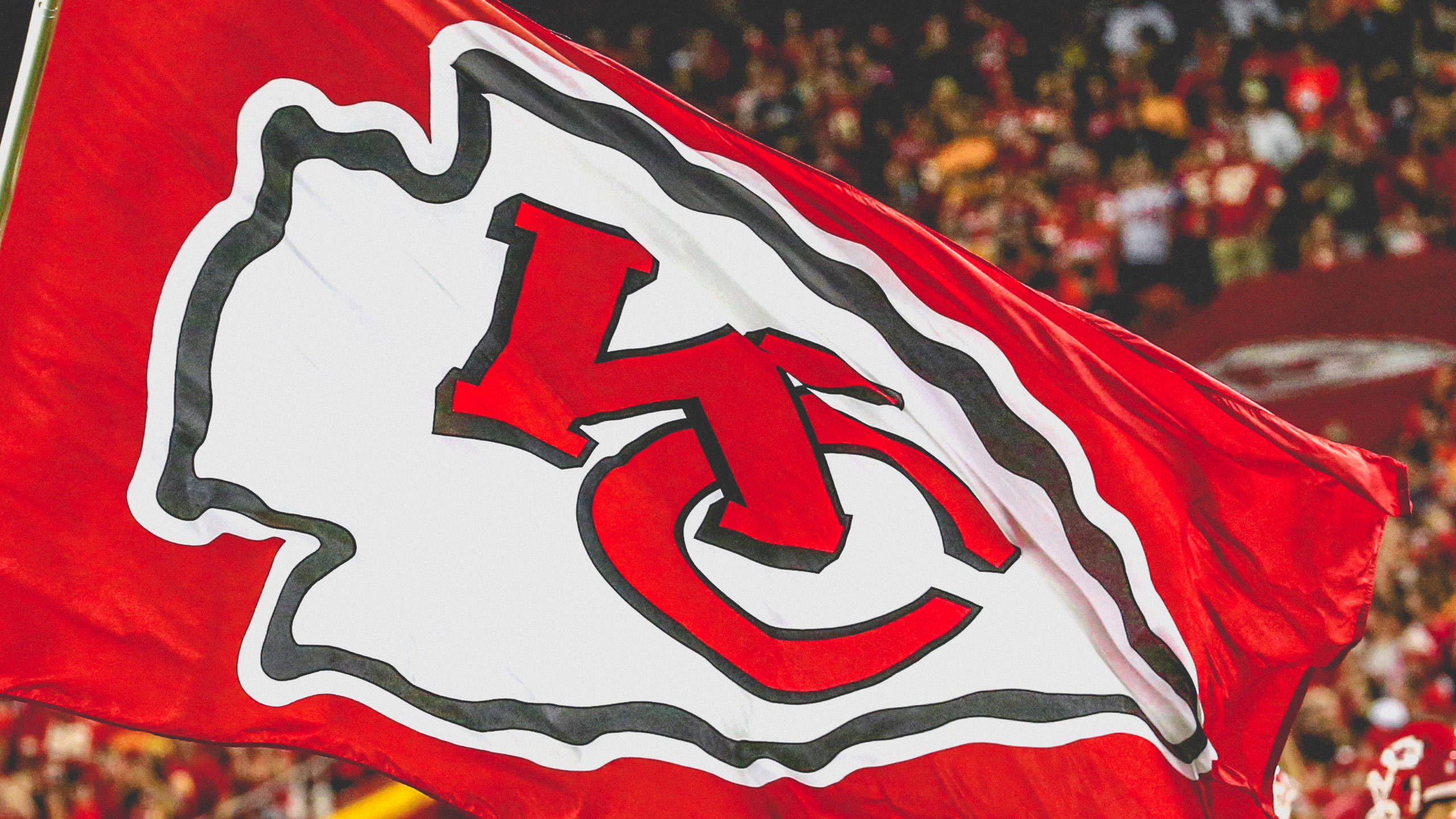Chiefs Helmet 桌布 壁紙 — 一面帶有白色和黑色標誌的紅色堪薩斯城酋長旗幟，在模糊的體育場人群前飄揚。