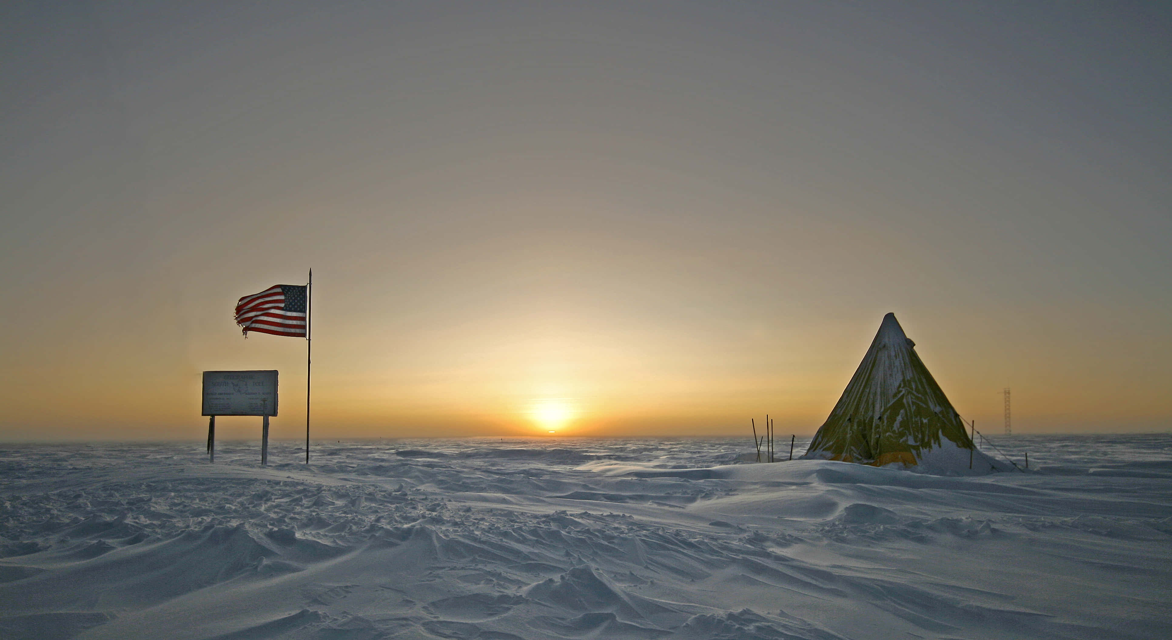 South Pole 桌布 壁紙 — 一面廣闊的冰凍平原上，一面孤獨的美國國旗和一個被雪覆蓋的帳篷矗立著，夕陽在地平線上緩緩落下。