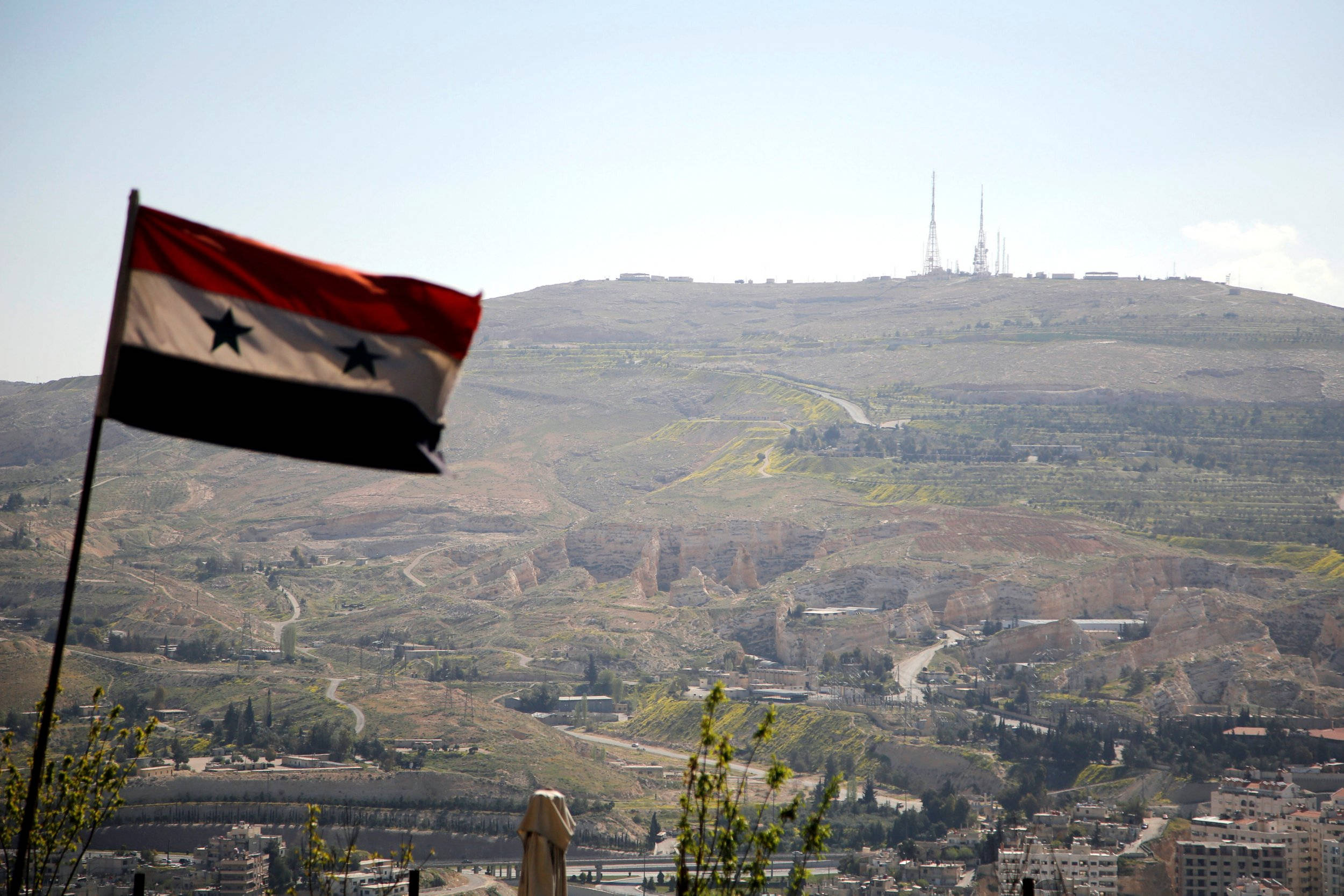 Syria 桌布 壁紙 — 一面敘利亞國旗在前景飄揚，背景是廣闊的丘陵地帶，遠處山頂上有通訊塔。