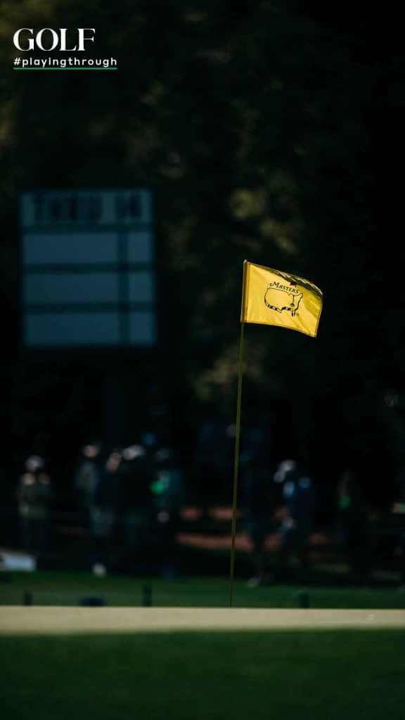 Augusta National iPhone 桌布 壁紙 — 一面明亮的黃色大師賽高爾夫旗幟在微風中飄揚，背景是昏暗的果嶺和模糊的觀眾。