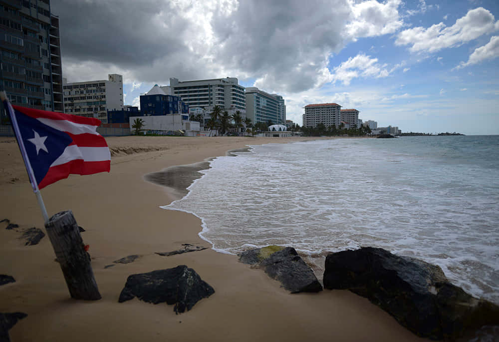 Puerto Rican 桌布 壁紙 — 一面波多黎各國旗在沙灘上飄揚，海浪拍打岩石，一排酒店在陰天之下。