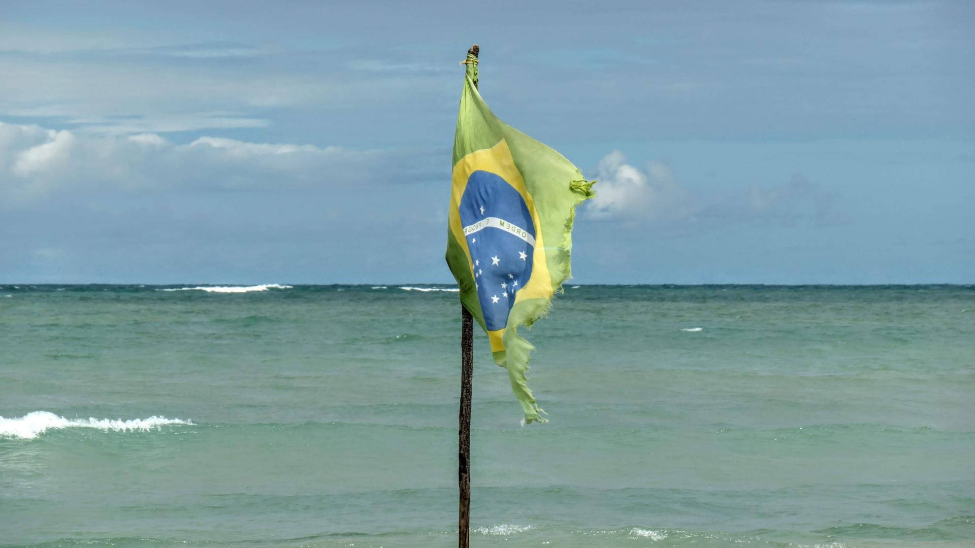 Brazil Flag 桌布 壁紙 — 一面破爛的巴西國旗在木桿上飄揚，背景是海浪和部分多雲的天空。