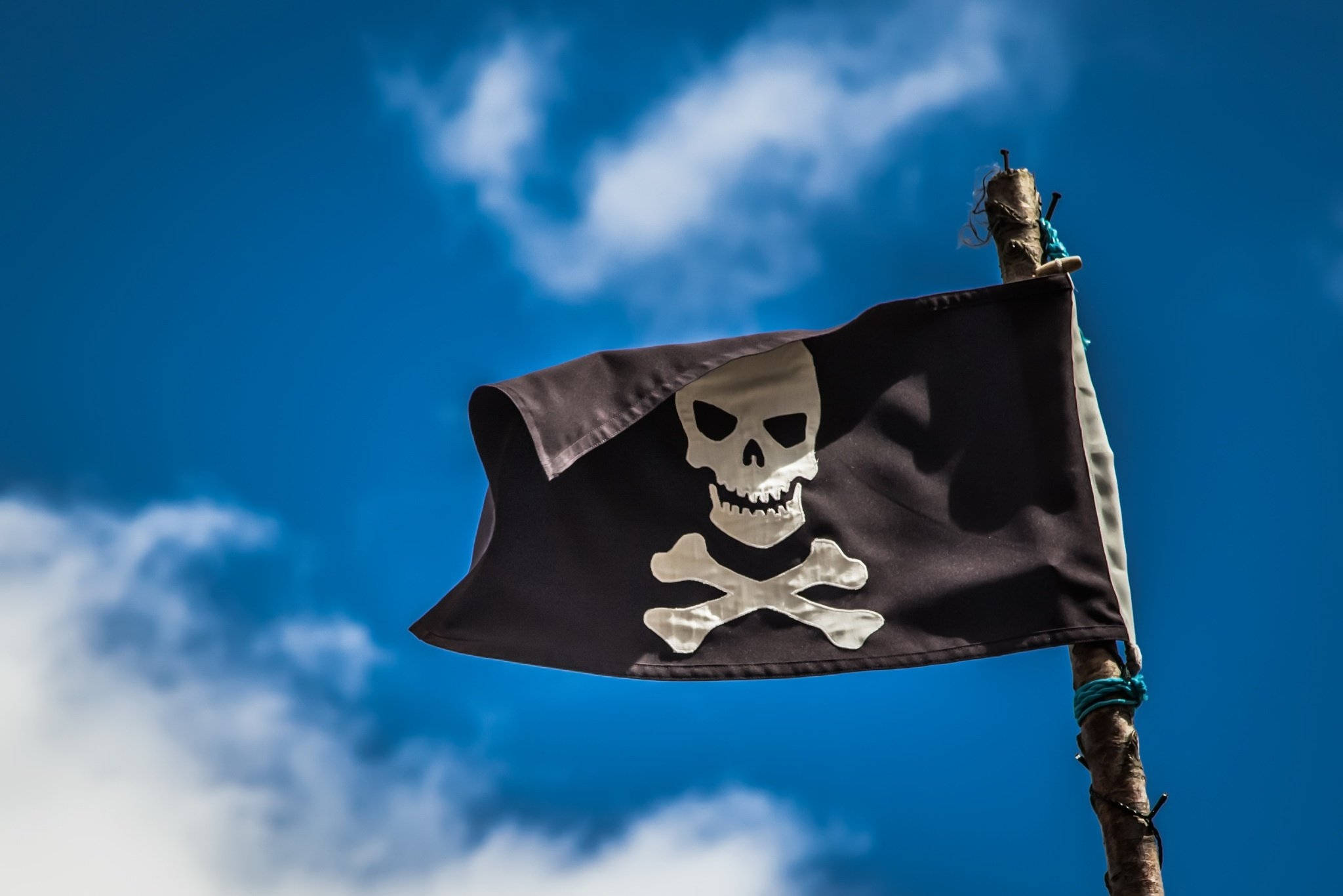 Flag 桌布 壁紙 — 一面破爛的黑色海盜旗，上面有白色骷髏頭和交叉骨頭，在藍天白雲中飄揚。