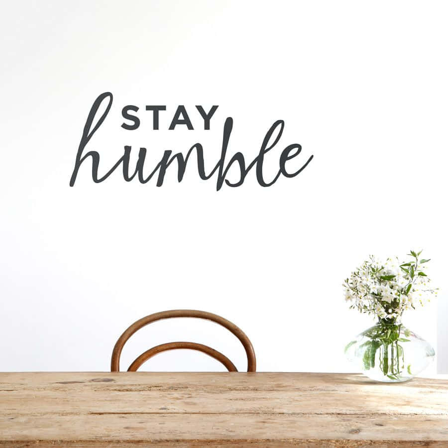 語錄 桌布 壁紙 — 一面簡約白色牆壁上，黑色字體寫著「STAY humble」，下方有一張木桌，桌上放著一瓶白色花和一把椅子。