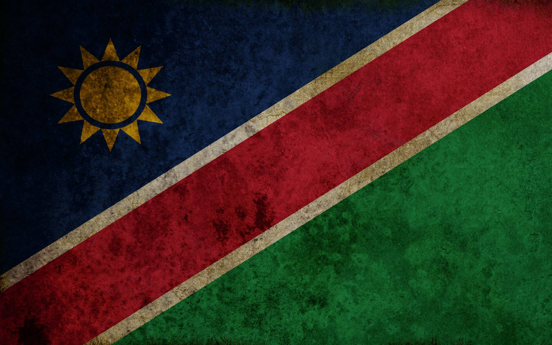 Namibia 桌布 壁紙 — 一面經風化的納米比亞國旗，帶有藍色、紅色和綠色區域以及黃色太陽標誌，顯示出可見的紋理和磨損。
