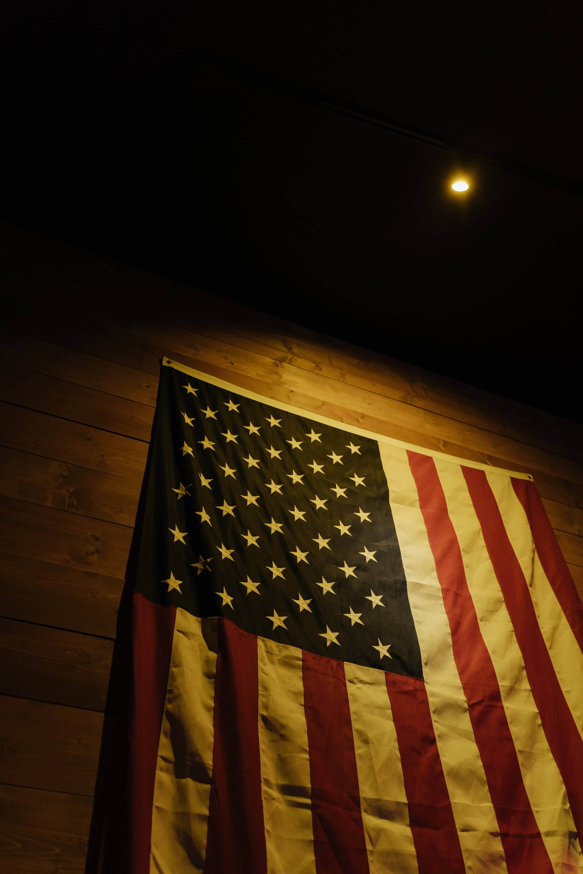 Black American Flag 桌布 壁紙 — 一面美國國旗掛在木牆上，在昏暗的房間裡被一盞溫暖的聚光燈照亮。