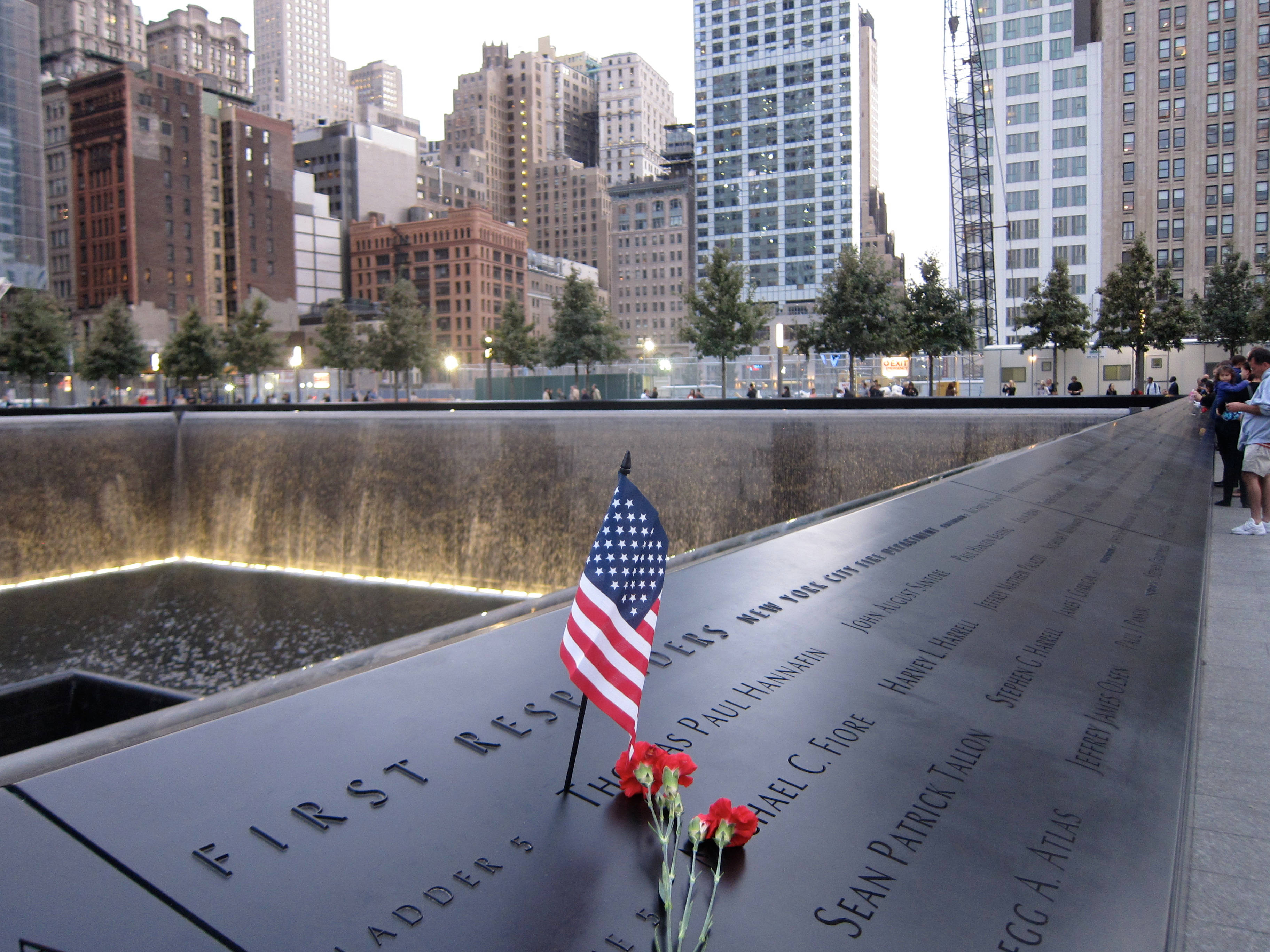 911 Memorial 桌布 壁紙 — 一面莊嚴的美國國旗和紅色鮮花安放在911紀念館的倒影池邊，周圍是高聳的城市建築。