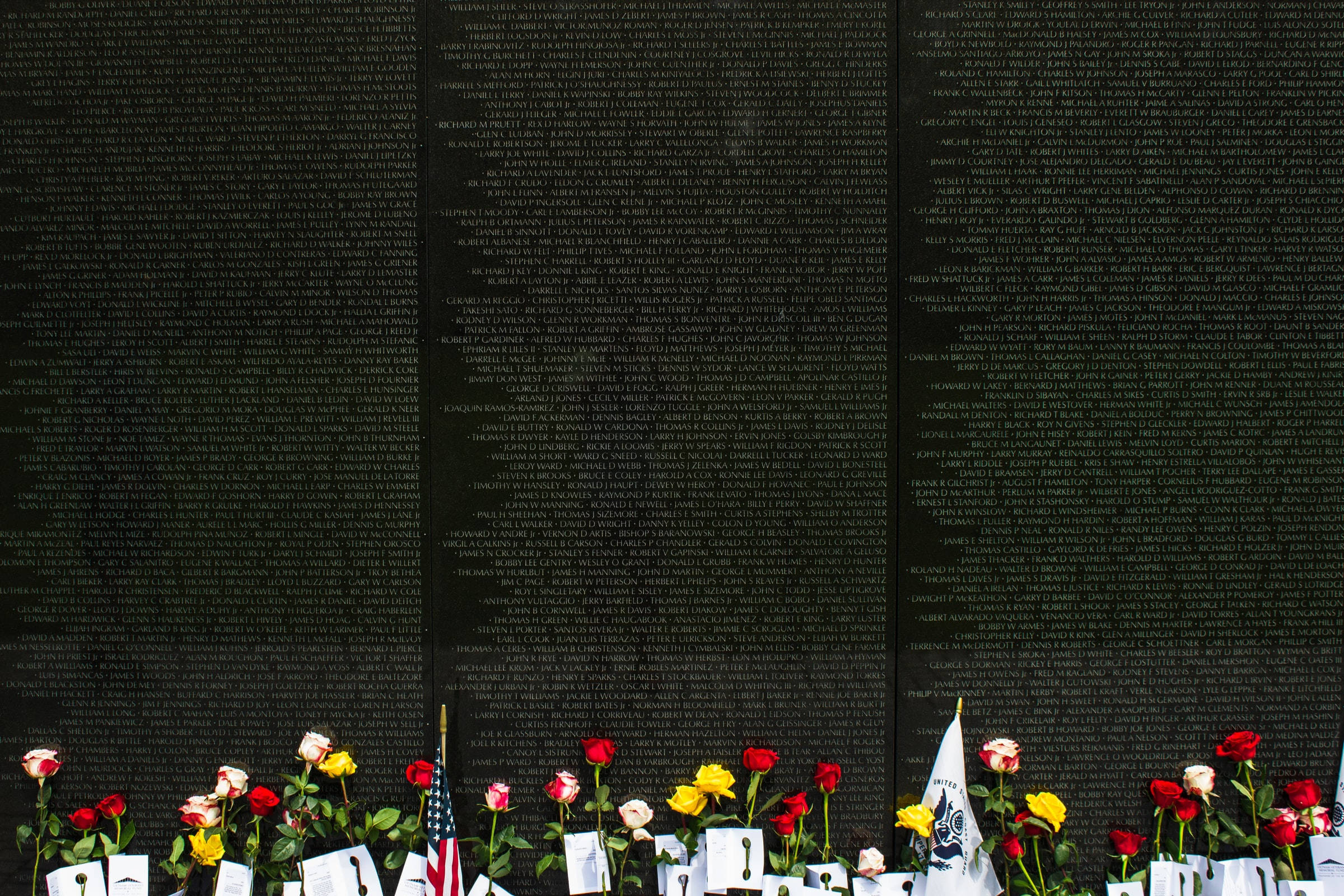 Remembrance 桌布 壁紙 — 一面莊嚴的黑色紀念牆，刻有名字，底部裝飾著紅、黃、白色的玫瑰和小型美國國旗。