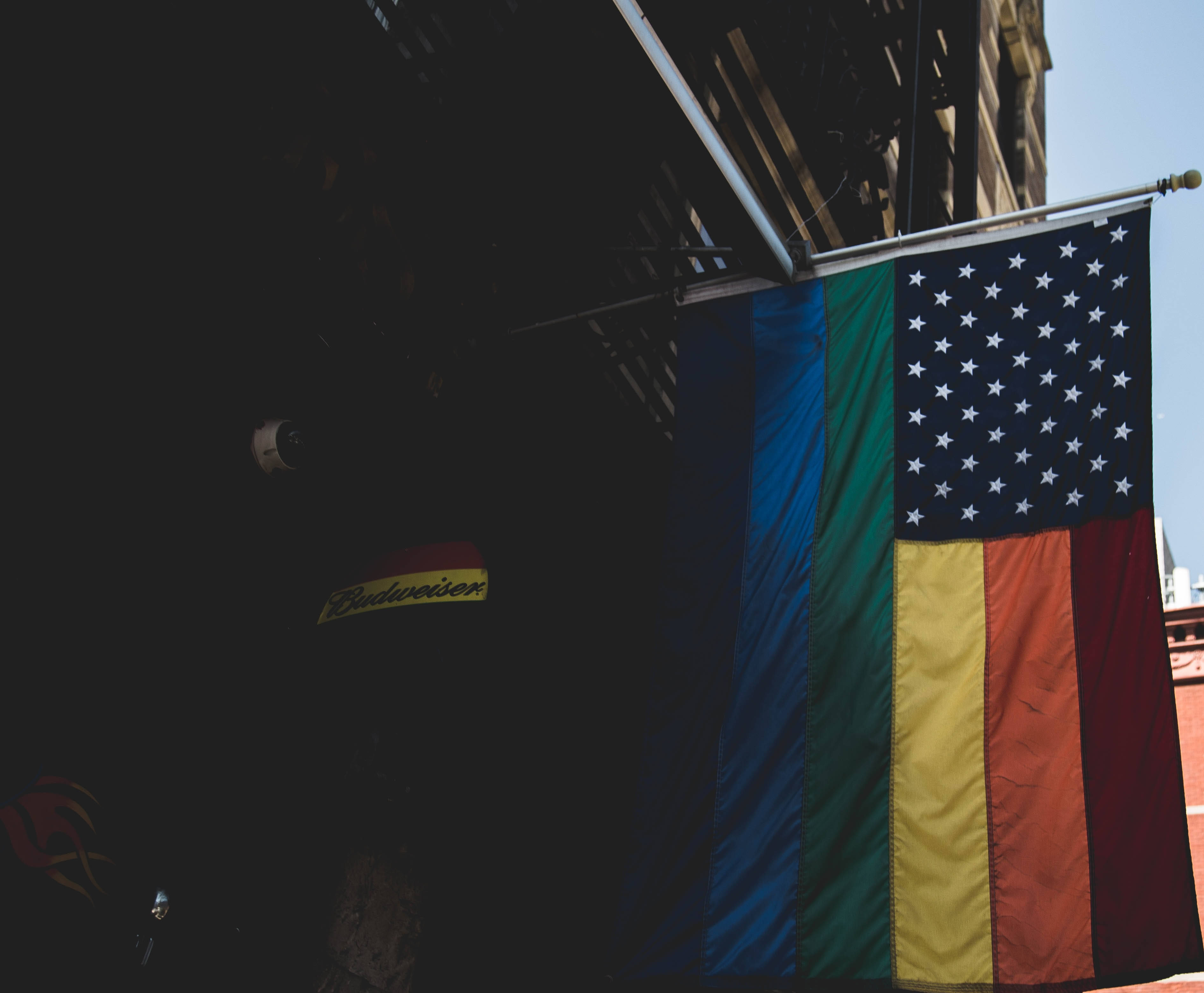Pride Flag 桌布 壁紙 — 一面融合了美國國旗的驕傲旗幟掛在建築物的遮陽篷上，背景是明亮的藍天。