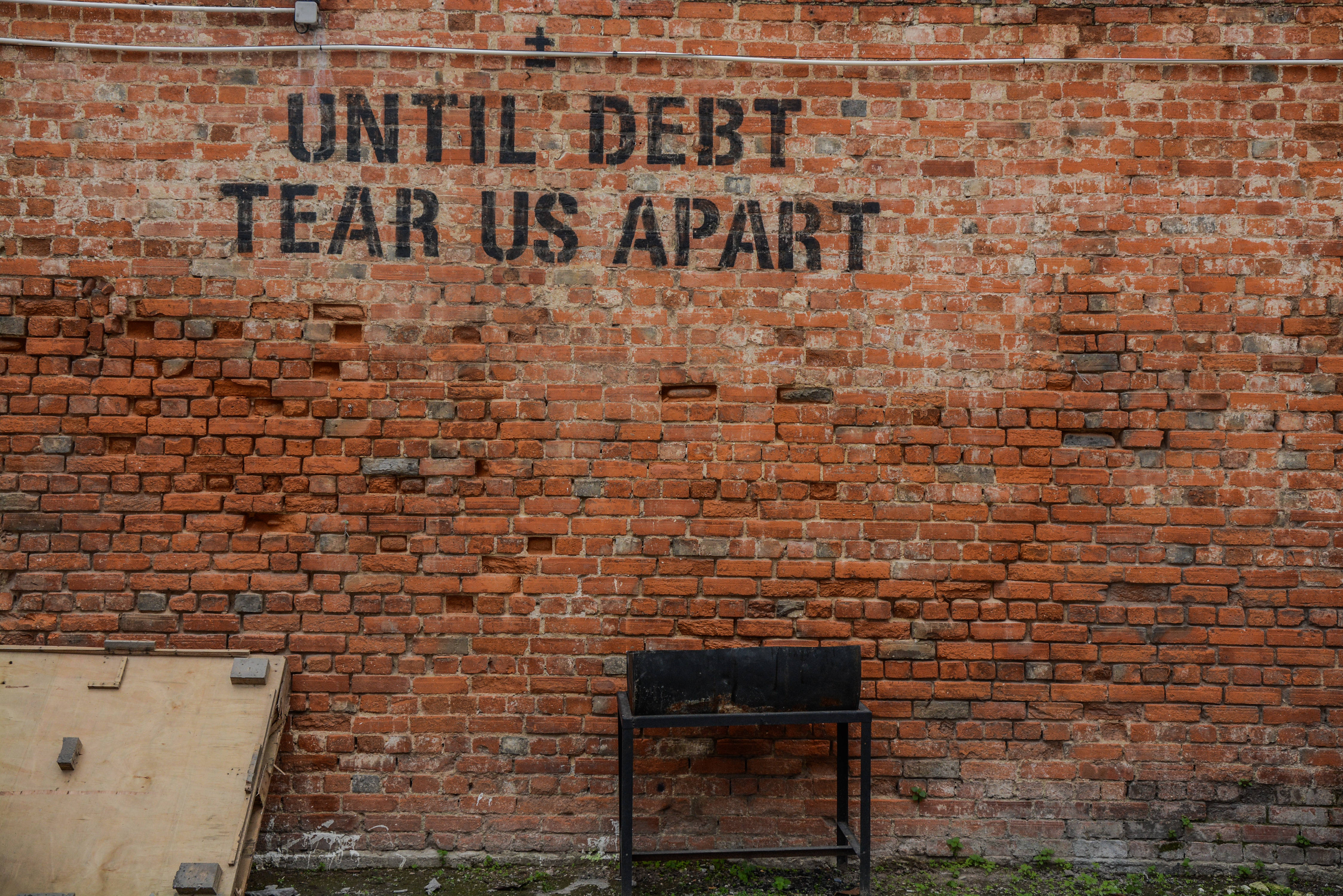 Accounting 桌布 壁紙 — 一面風化的磚牆上，有粗體黑色塗鴉寫著「UNTIL DEBT TEAR US APART」，上方是一張孤獨的金屬長椅。