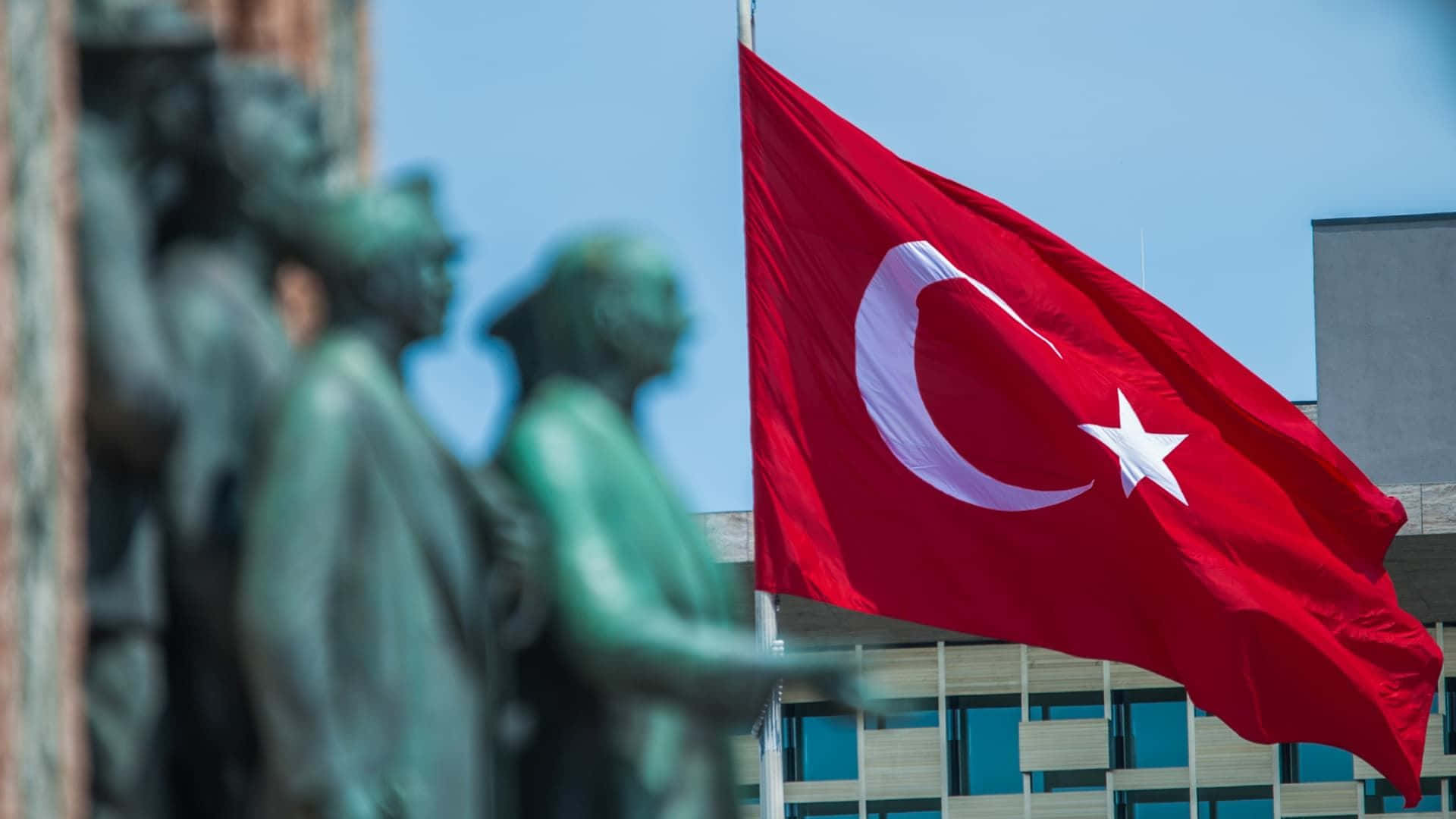 Turkish 桌布 壁紙 — 一面鮮紅的土耳其國旗，帶有白色新月和星星，在晴朗的藍天下隨風飄揚，前景有模糊的青銅雕像。