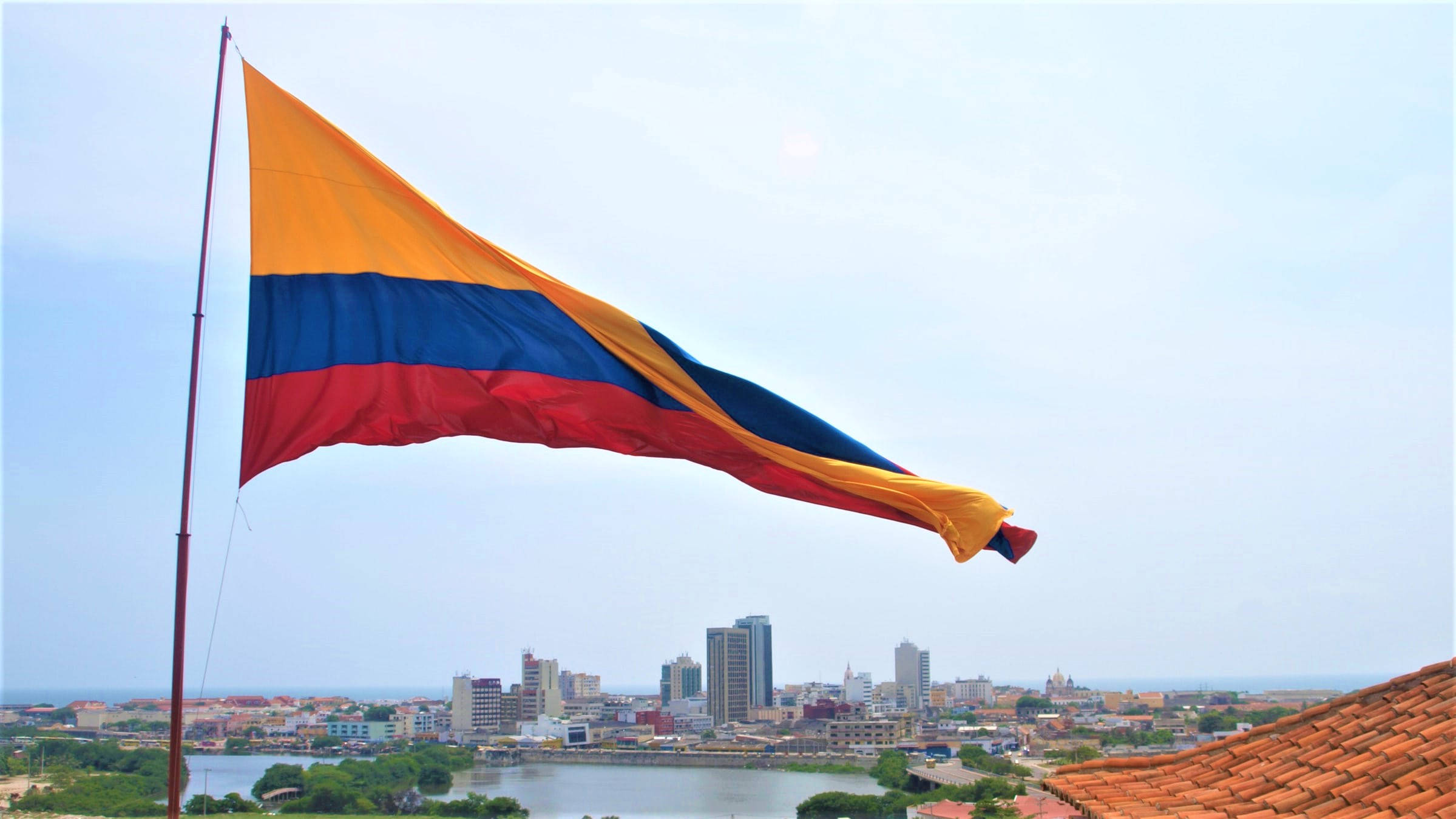 Colombia Flag 桌布 壁紙 — 一面鮮豔的哥倫比亞國旗在旗桿頂端驕傲飄揚，背景是朦朧的天空，俯瞰著擁有河流和瓦片屋頂的廣闊城市景觀。