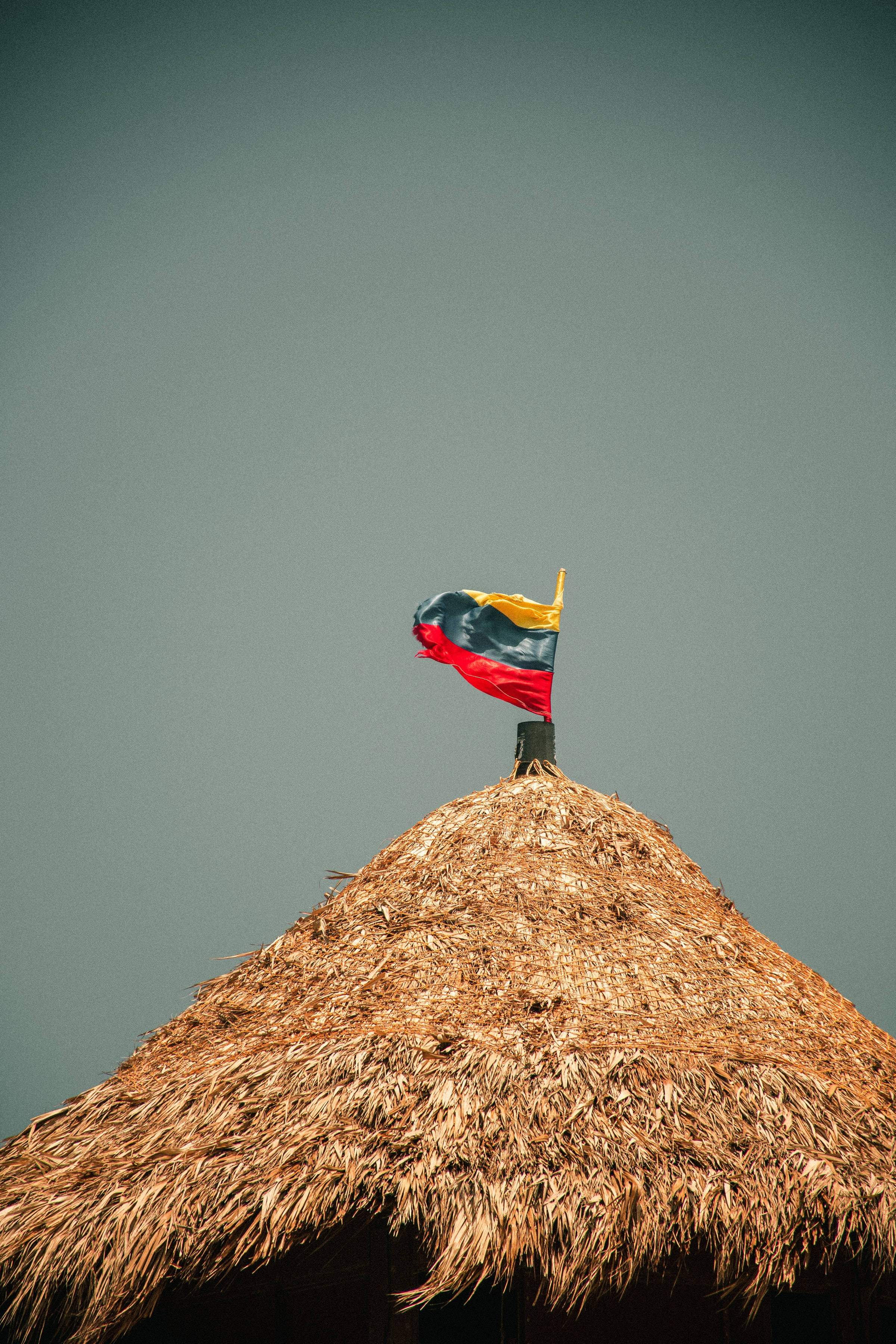 Colombia Flag 桌布 壁紙 — 一面鮮豔的哥倫比亞國旗在質樸的茅草屋頂上飄揚，背景是柔和的灰色天空。