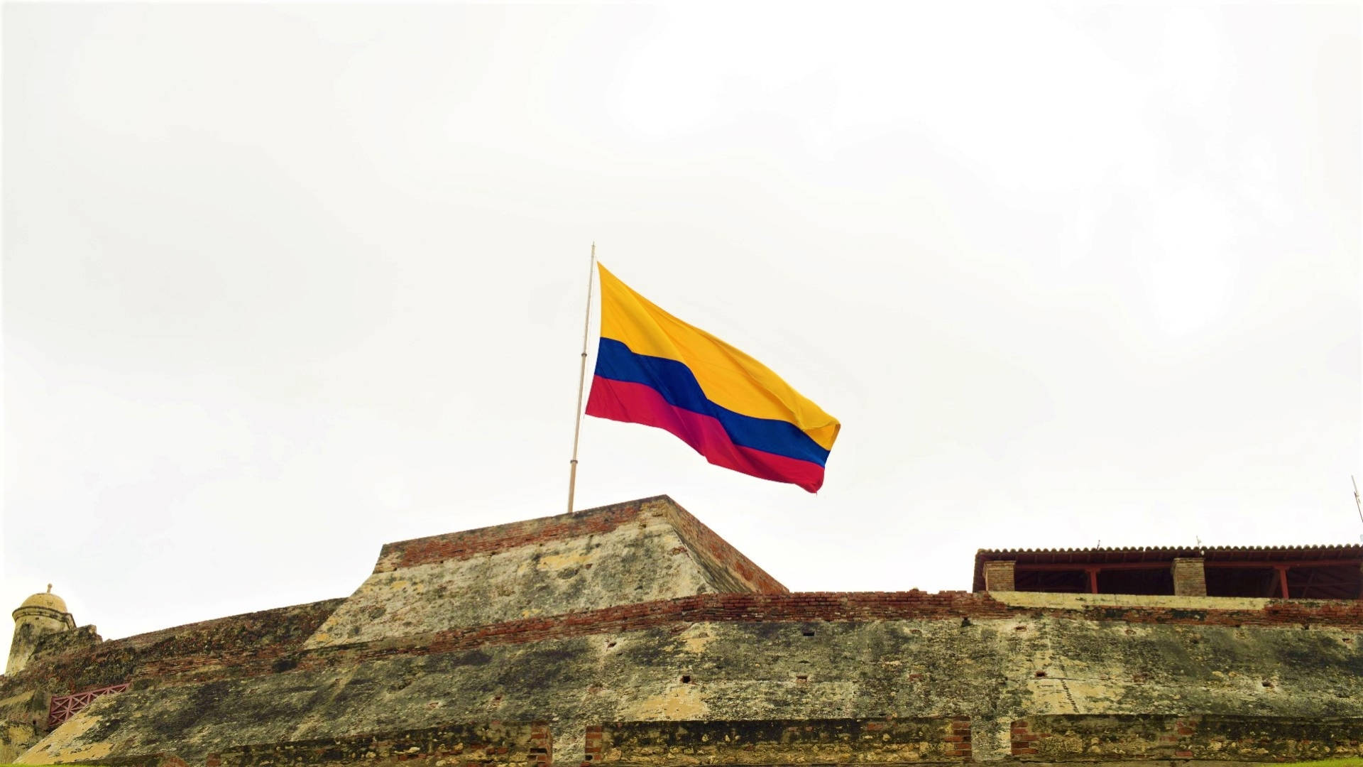 Colombia Flag 桌布 壁紙 — 一面鮮豔的哥倫比亞國旗在陰天之下，驕傲地飄揚在風化的石頭堡壘頂上。