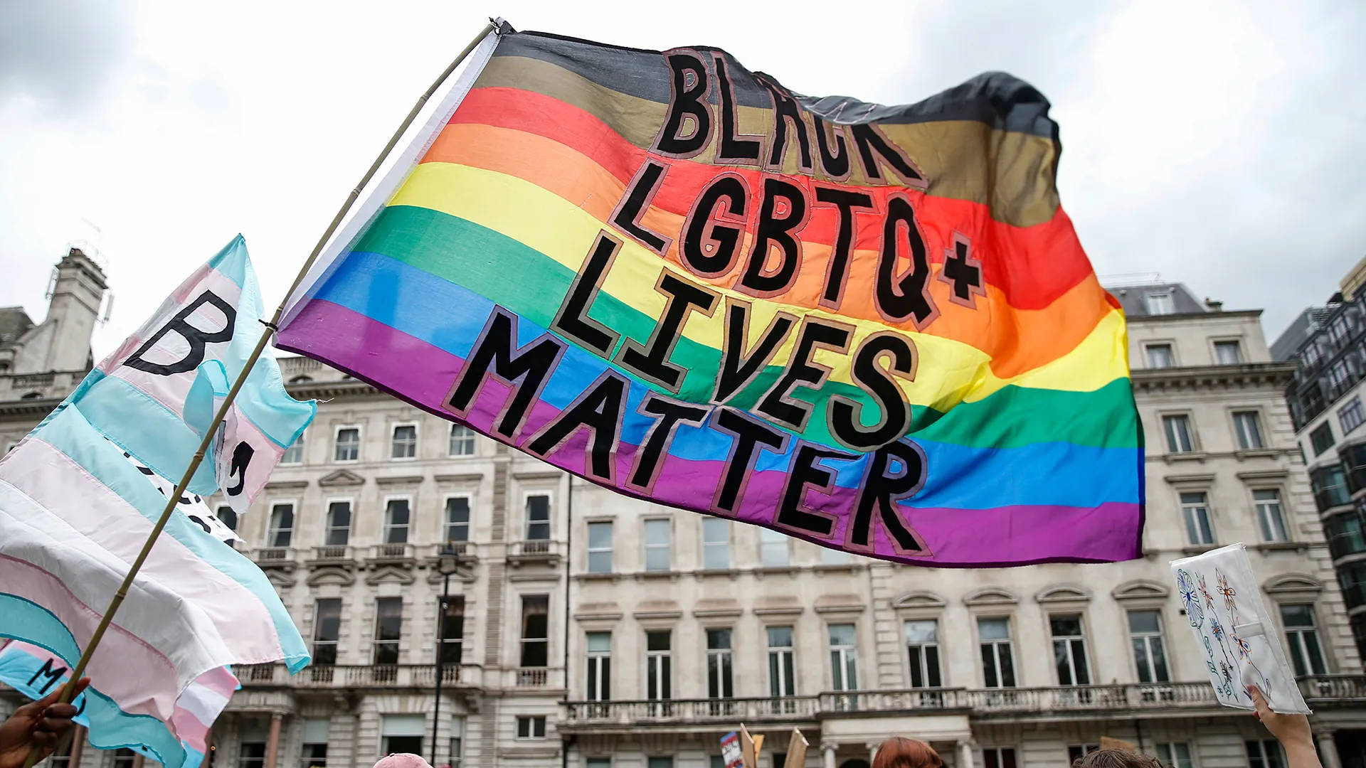 黑色 桌布 壁紙 — 一面鮮豔的彩虹旗，上面有粗體黑色文字寫著「BLACK LGBTQ+ LIVES MATTER」，在歷史悠久的城市建築背景下，於抗議活動中自豪地飄揚。