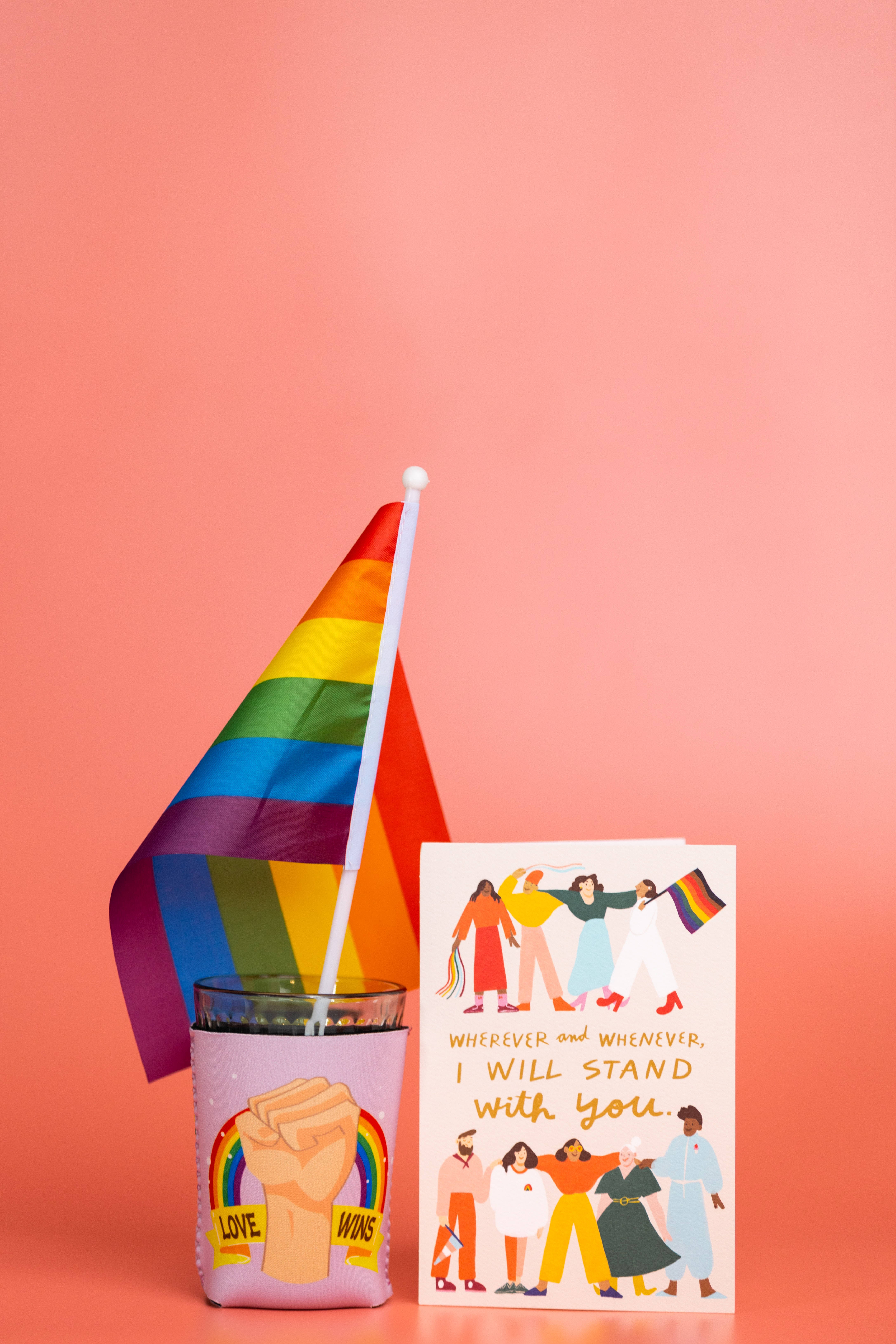 Pride Flag 桌布 壁紙 — 一面鮮豔的彩虹旗和團結卡，上面有不同的人物，旁邊是一個帶有舉起拳頭設計的玻璃杯，背景是柔和的珊瑚色。