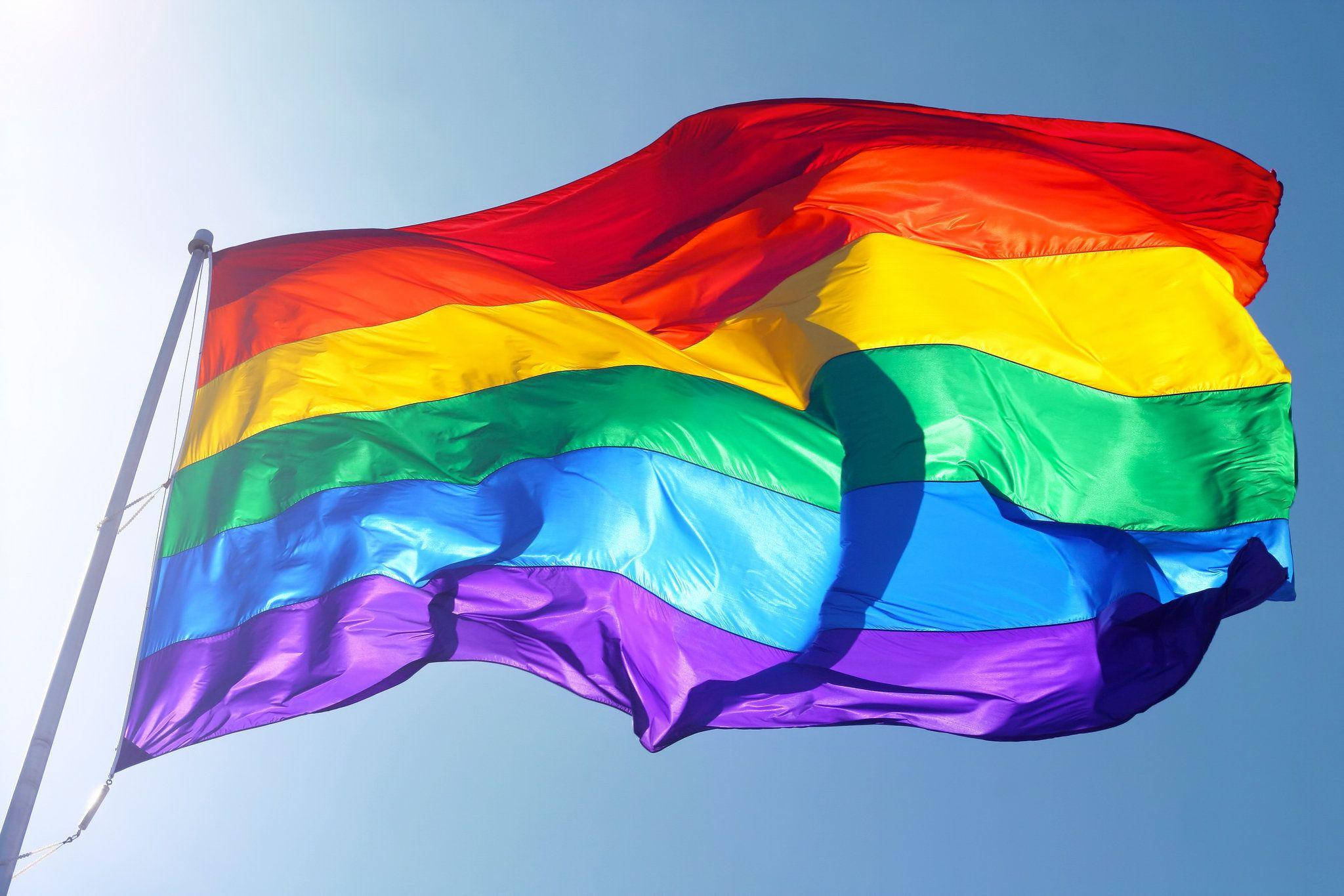 Pride Flag 桌布 壁紙 — 一面鮮豔的彩虹旗在明亮的陽光下，在清澈的藍天中動態飄揚。