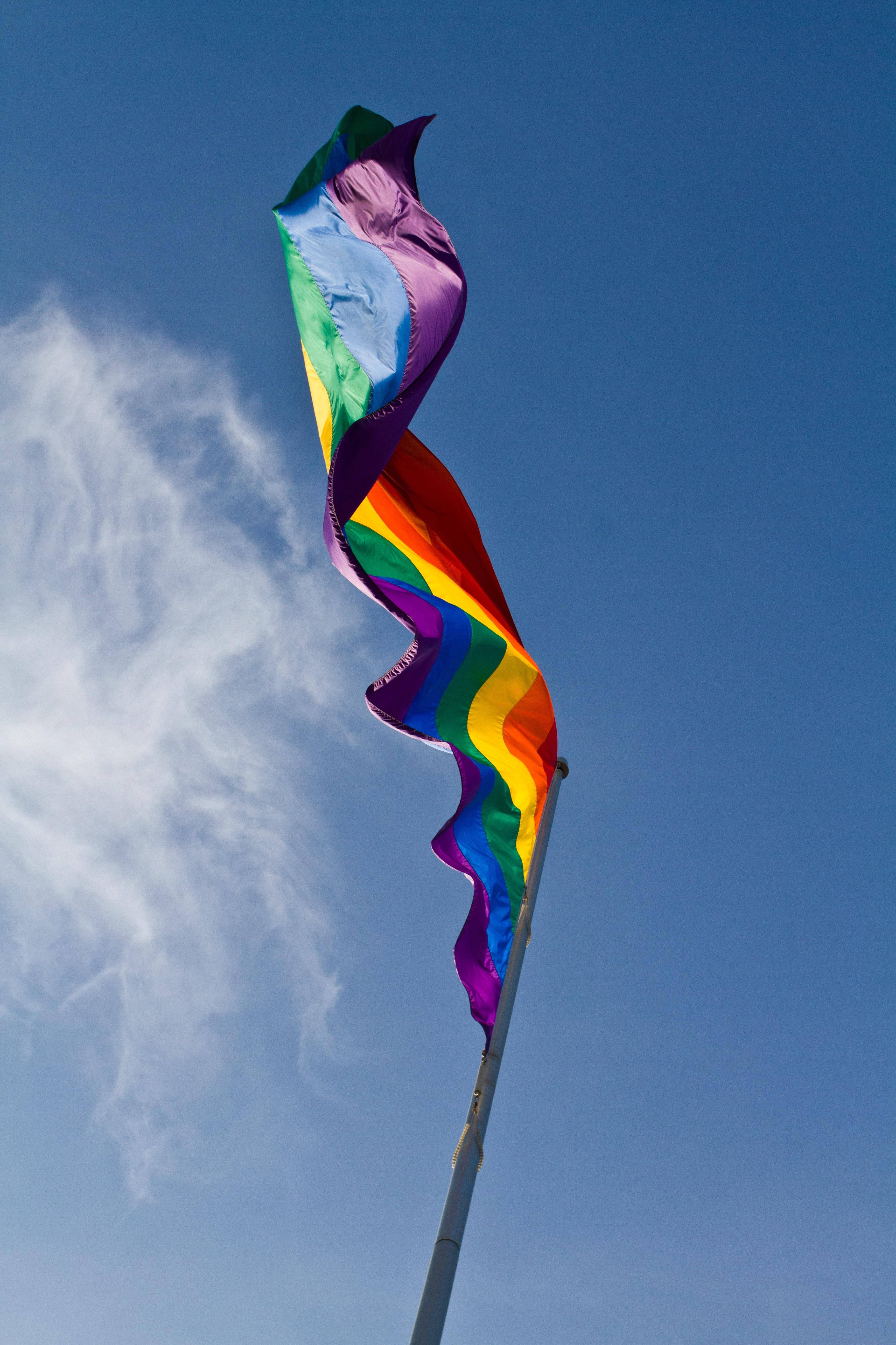 Pride Flag 桌布 壁紙 — 一面鮮豔的彩虹旗在晴朗藍天和縷縷白雲中動態飄揚。