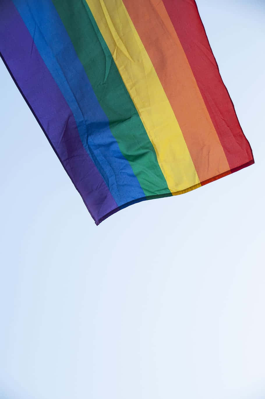 LGBT 桌布 壁紙 — 一面鮮豔的彩虹旗在柔和淡藍的天空中飄揚，輕柔的褶皺捕捉著光線。