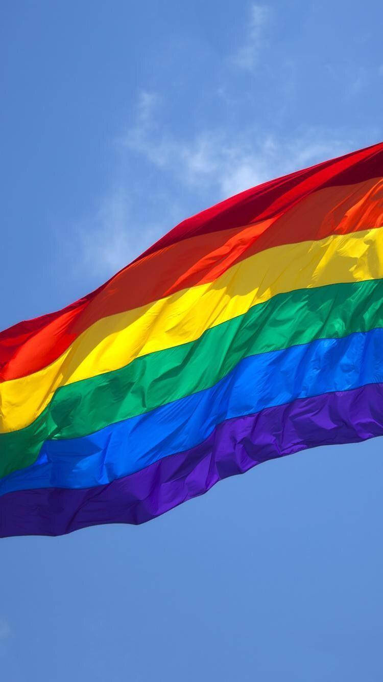 Pride Flag 桌布 壁紙 — 一面鮮豔的彩虹旗在藍天白雲下動態飄揚。