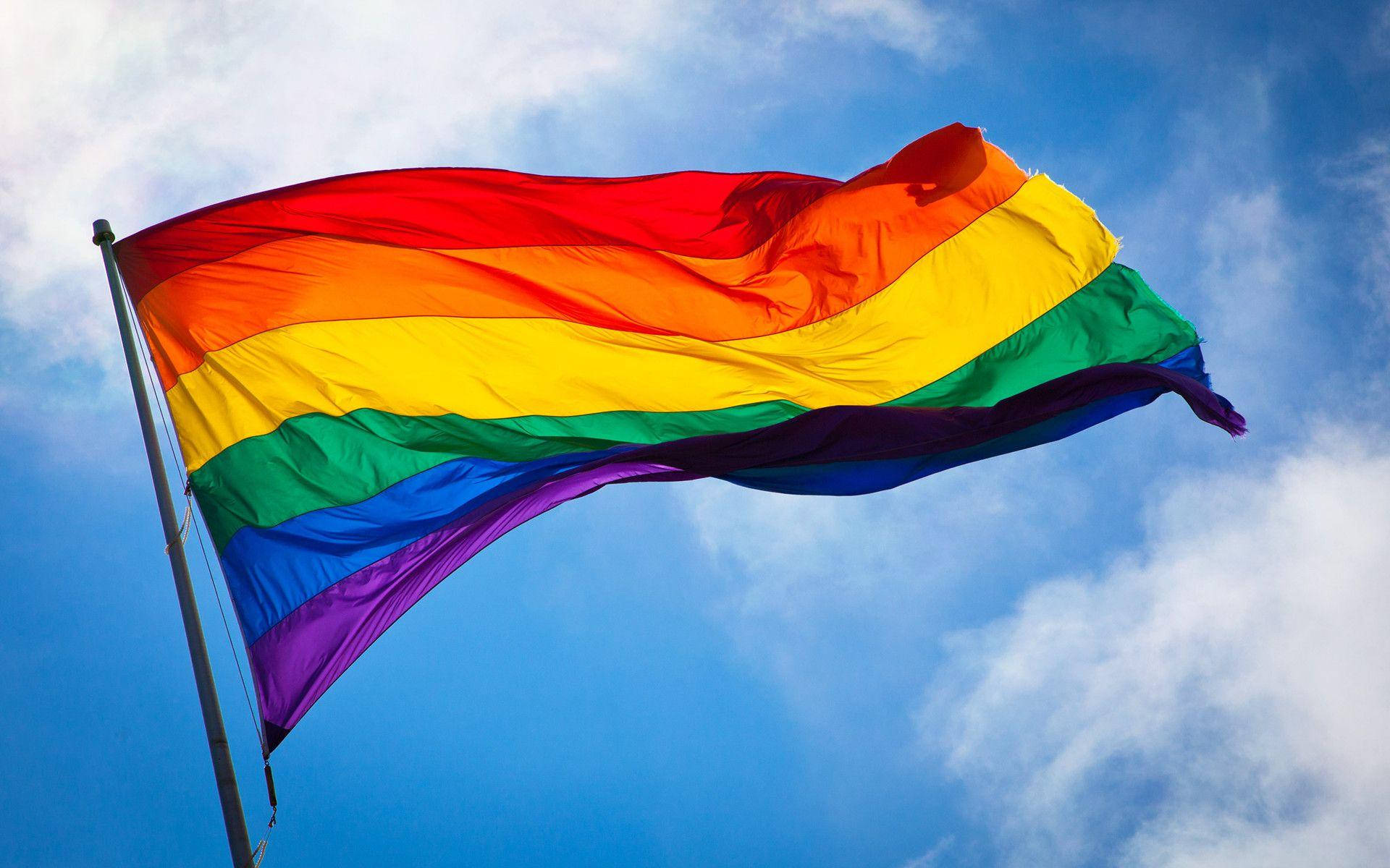 Pride Flag 桌布 壁紙 — 一面鮮豔的彩虹旗在藍天白雲中自豪地飄揚。