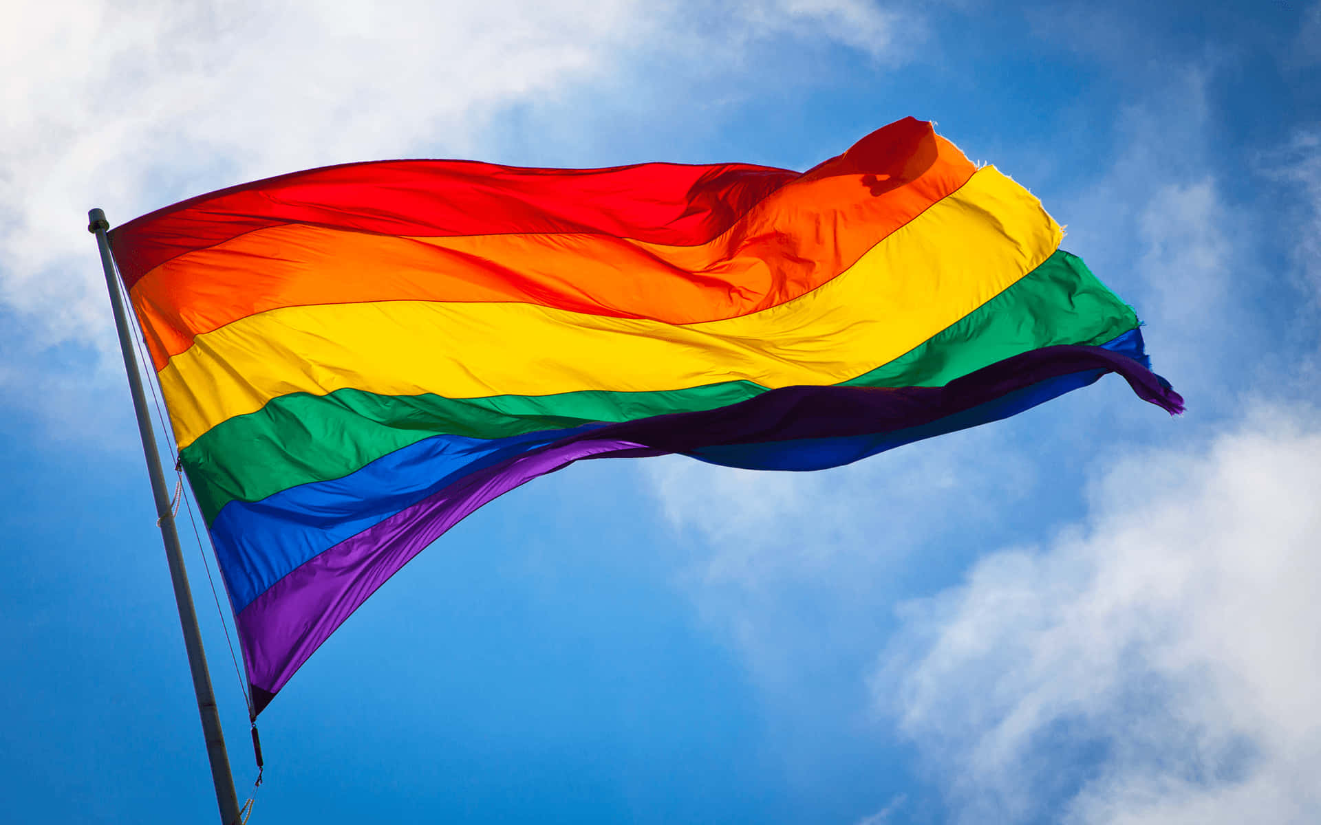 LGBT 桌布 壁紙 — 一面鮮豔的彩虹旗在點綴著 fluffy 白雲的明亮藍天下自豪地飄揚。