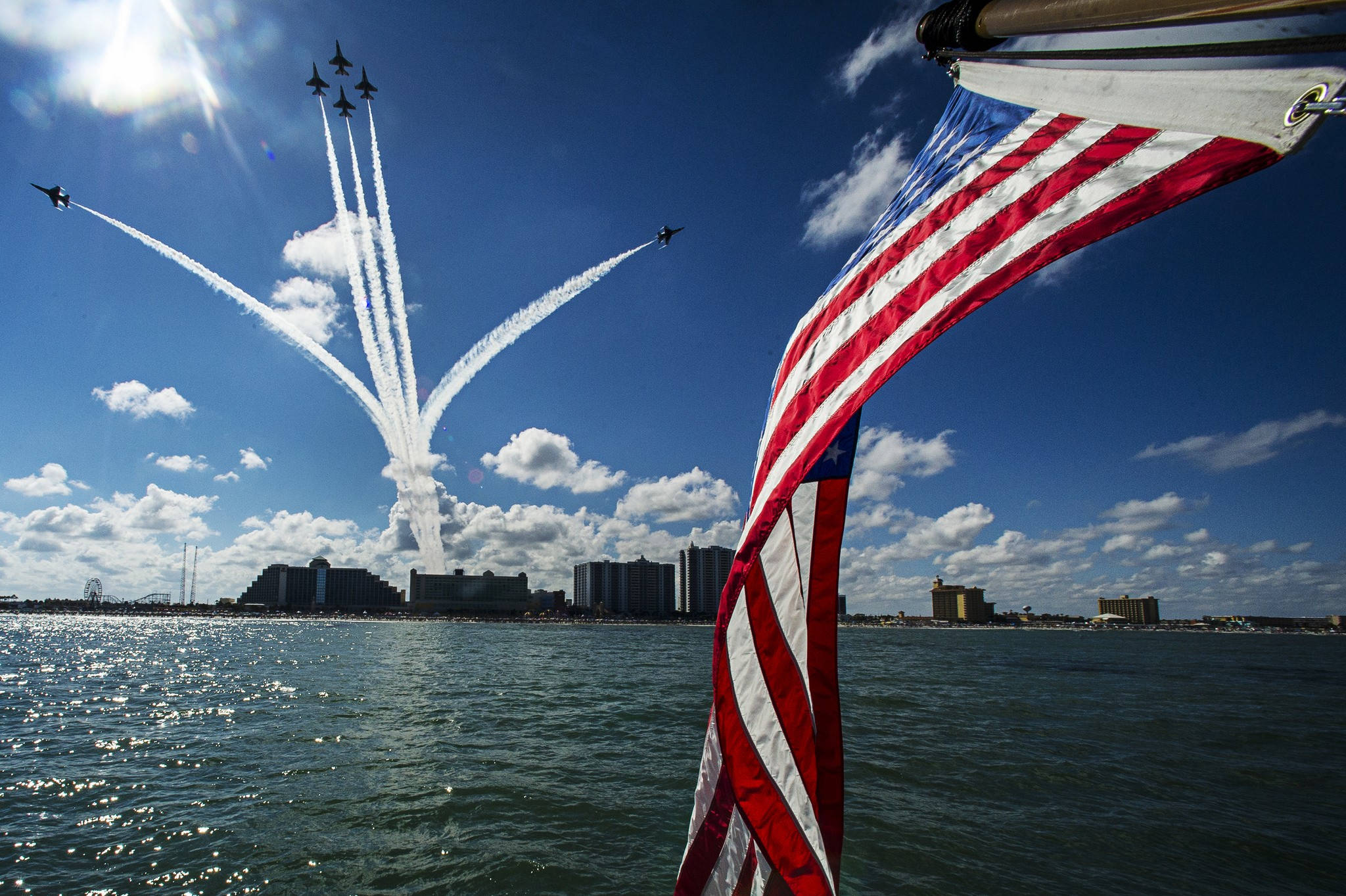 Military American Flag 桌布 壁紙 — 一面鮮豔的美國國旗在前景飄揚，戰鬥機在頭頂呼嘯而過，在明亮的藍天下，在閃閃發光的水濱城市上空留下白色煙跡。