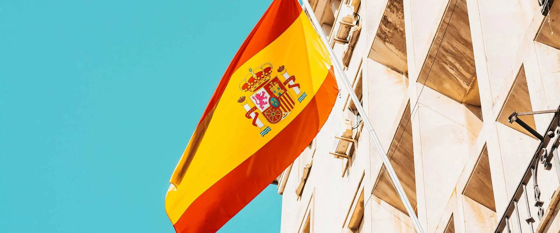 Spain Flag 桌布 壁紙 — 一面鮮豔的西班牙國旗在晴朗的藍天下，在幾何現代建築外牆旁飄揚。